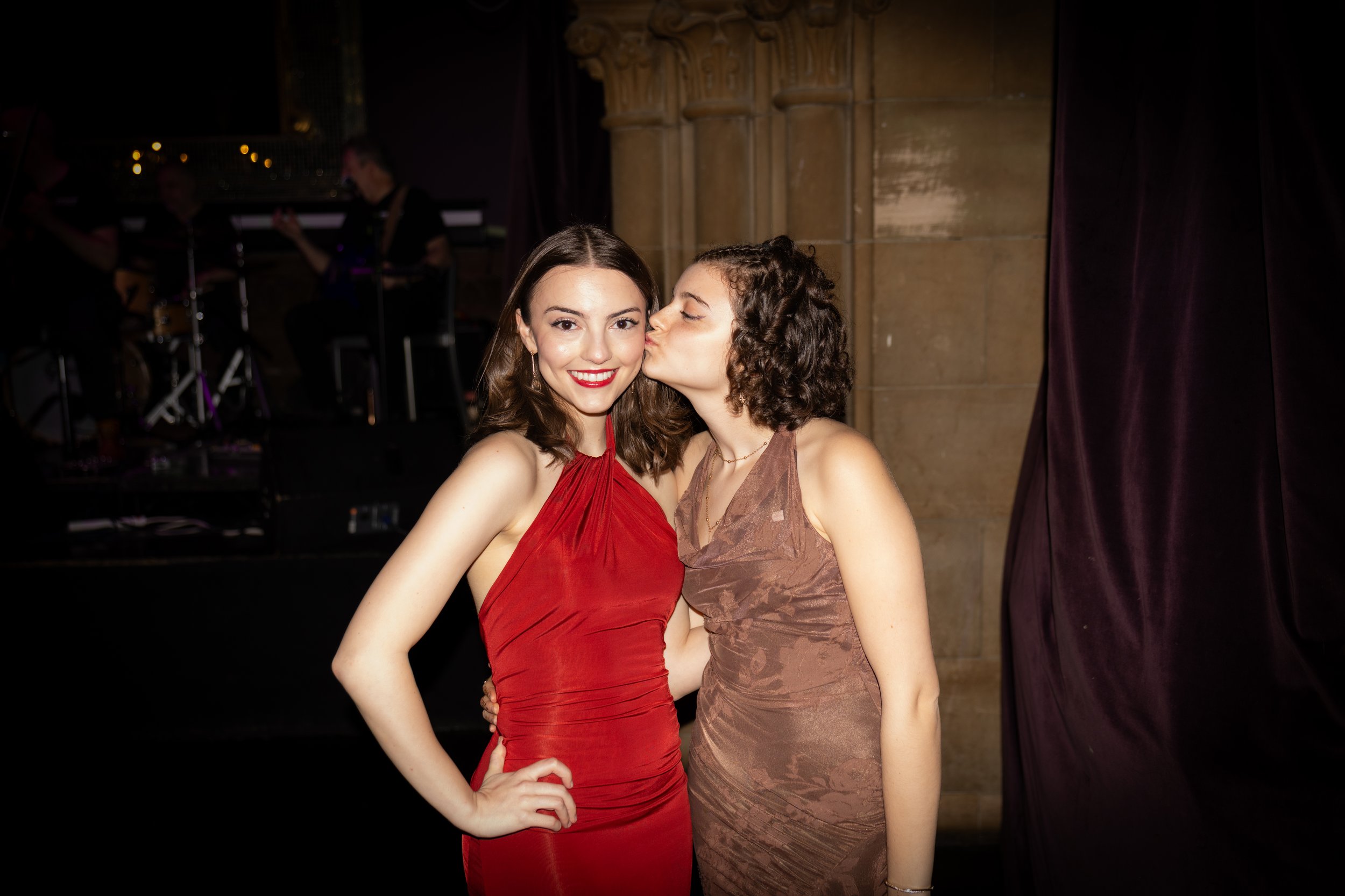 Two women in evening dresses, one in red and the other in brown, are standing close together. The woman in red is smiling at the camera while the woman in brown is giving her a kiss on the cheek. There are musicians in the background on a stage.