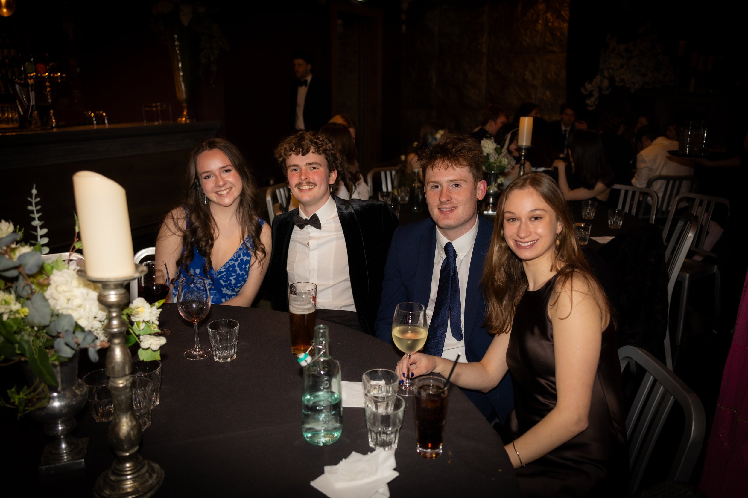 Four young adults, two women and two men, sit at a banquet table in a dimly lit venue, smiling at the camera. The table has glasses, a floral centerpiece, and candles.