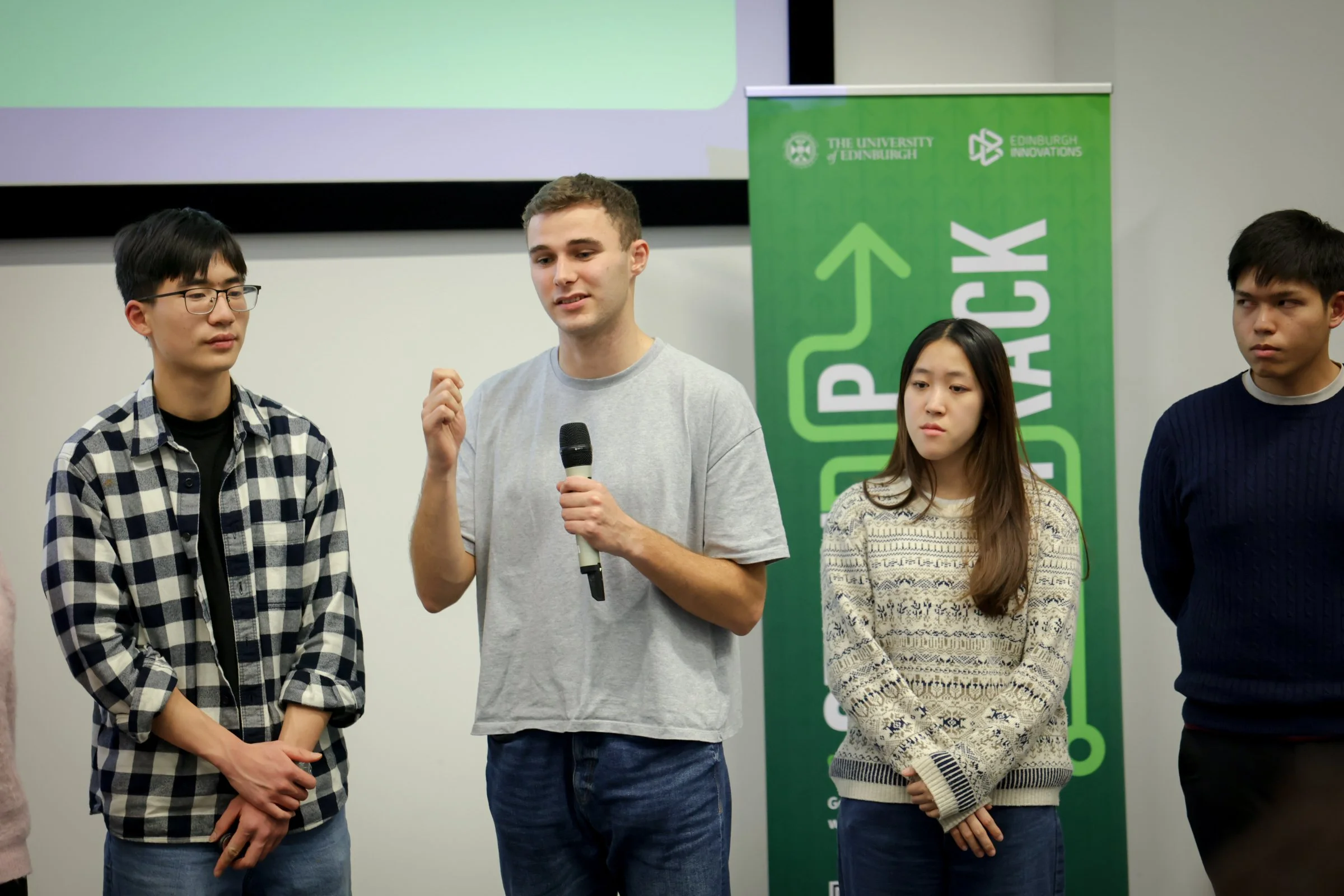 Group of four students standing on stage, one speaking into a microphone, with a green 'UP YOUR IMPACT' banner in the background at a university event.