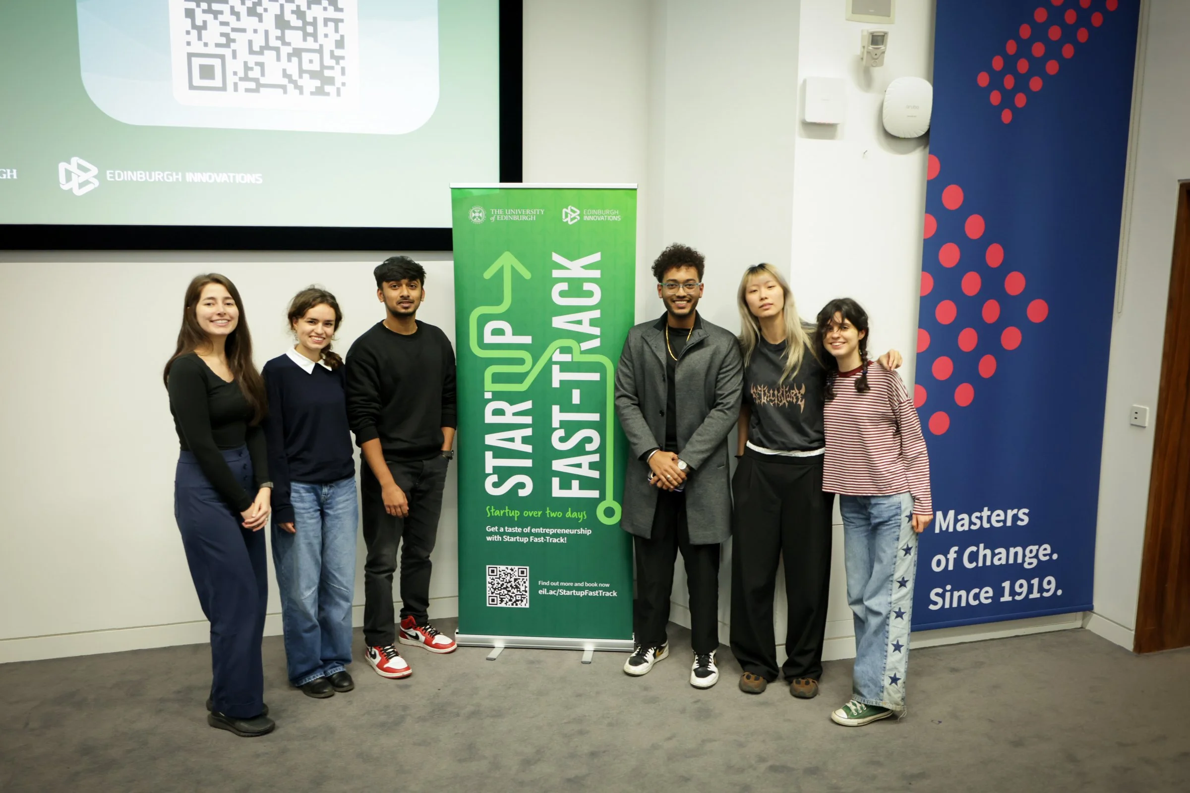 Group of six young adults standing indoors next to a green vertical banner promoting a startup fast-track event, and a blue banner with red dots and white text about a history of change since 1919. The group appears happy and casual, with some smilin