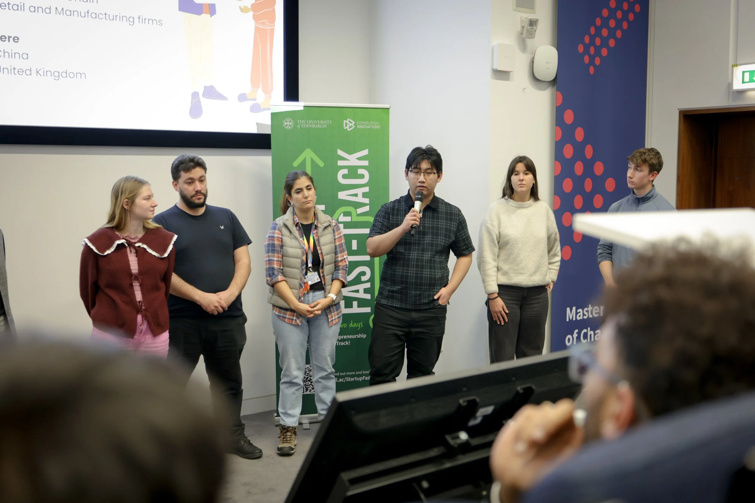 Group of six young adults standing in front of a presentation screen and banner at a panel discussion or startup event, with audience members visible in the foreground.