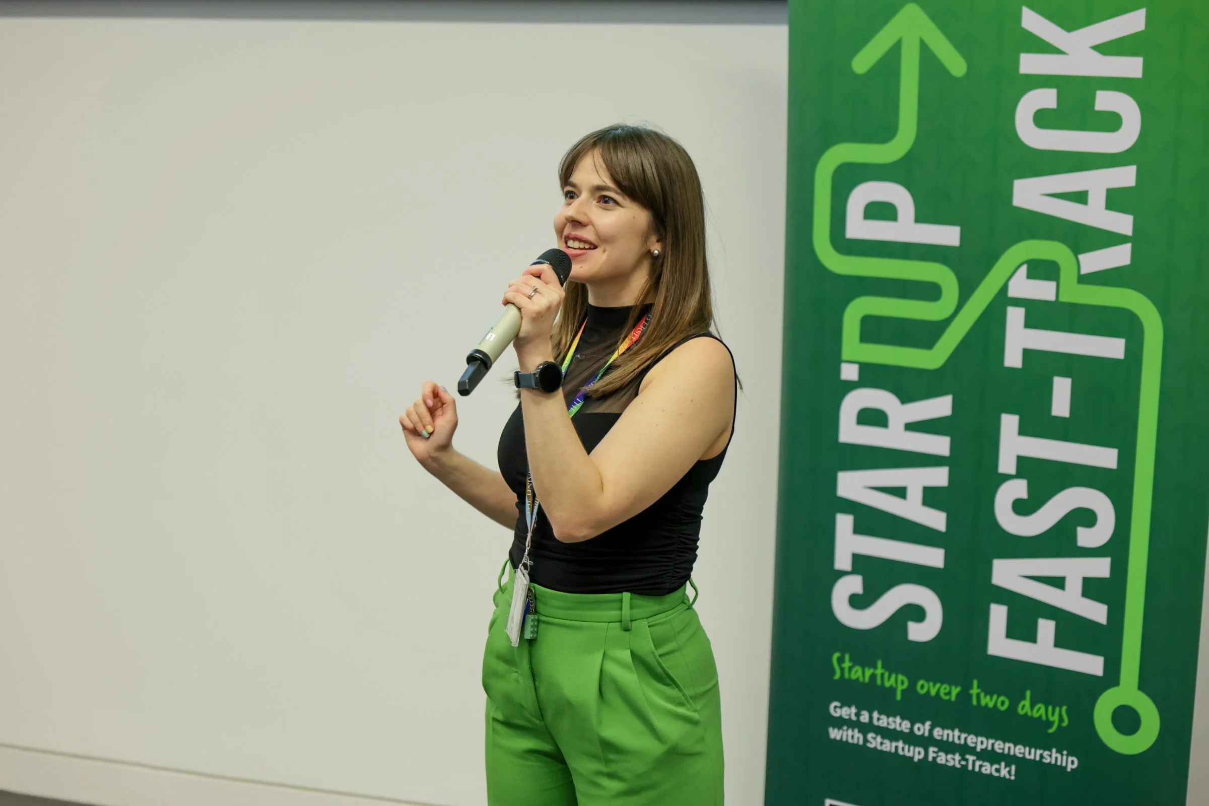 A woman holding a microphone and speaking at a startup event, standing beside a green banner that reads 'STARTUP FAST-TRACK' and 'Startup over two days. Get a taste of entrepreneurship with Startup Fast-Track!'
