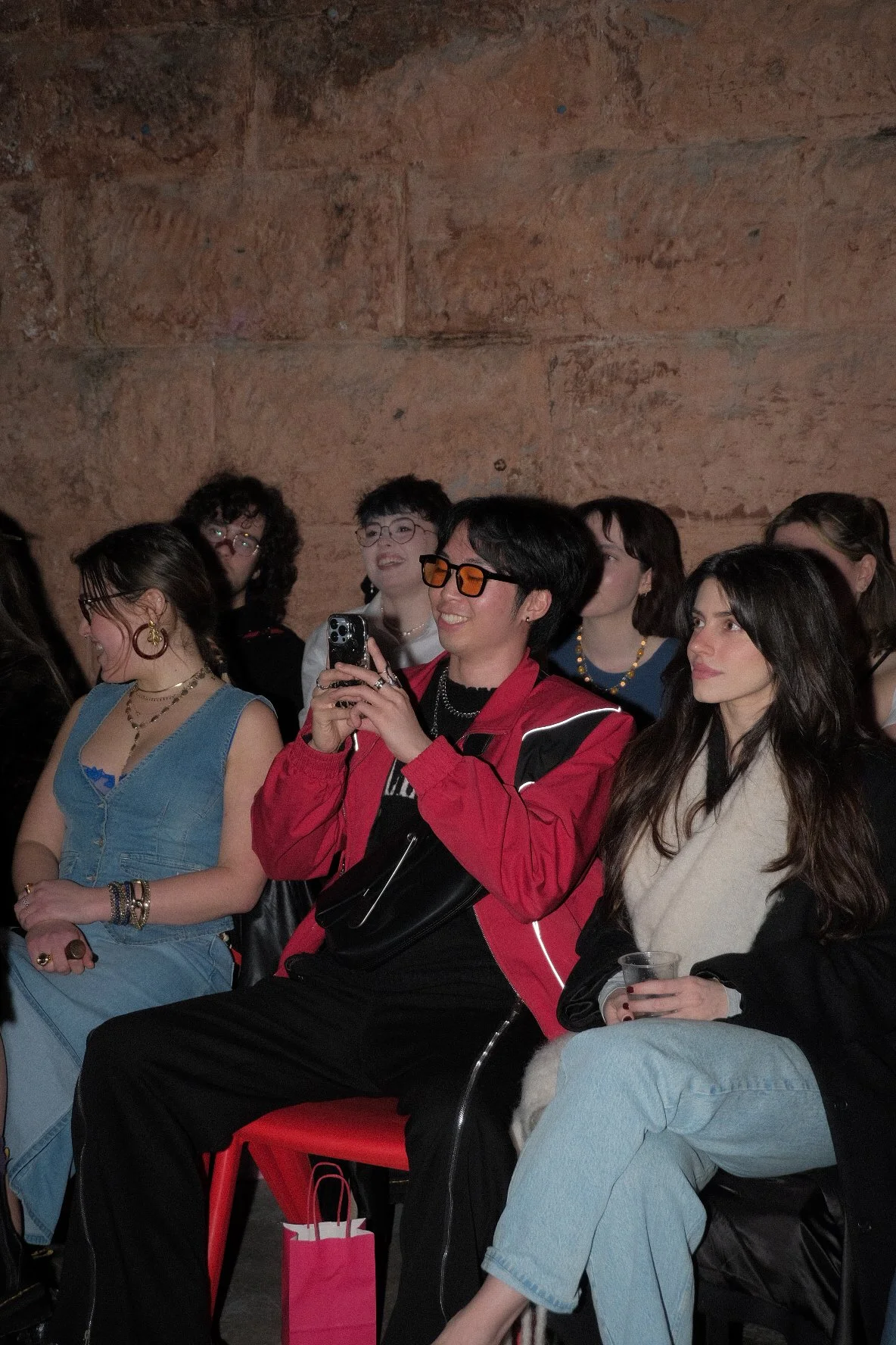 A group of people sitting and smiling at an indoor event with a brick wall background. One person in the center is taking a photo with a phone, wearing glasses and a red jacket.