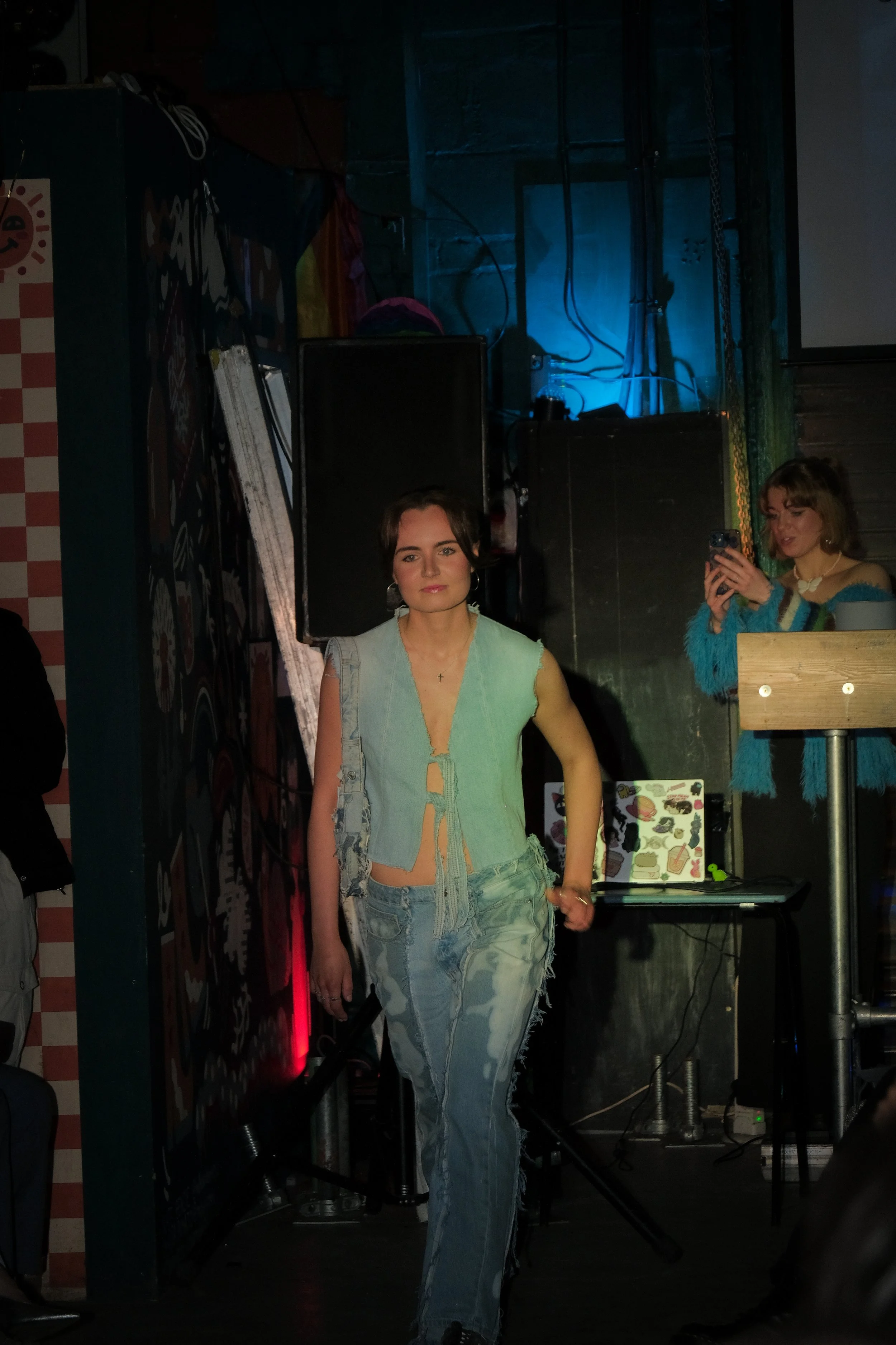 A young woman with short brown hair standing indoors, wearing a light denim vest and ripped jeans, with another woman in the background taking a photo.