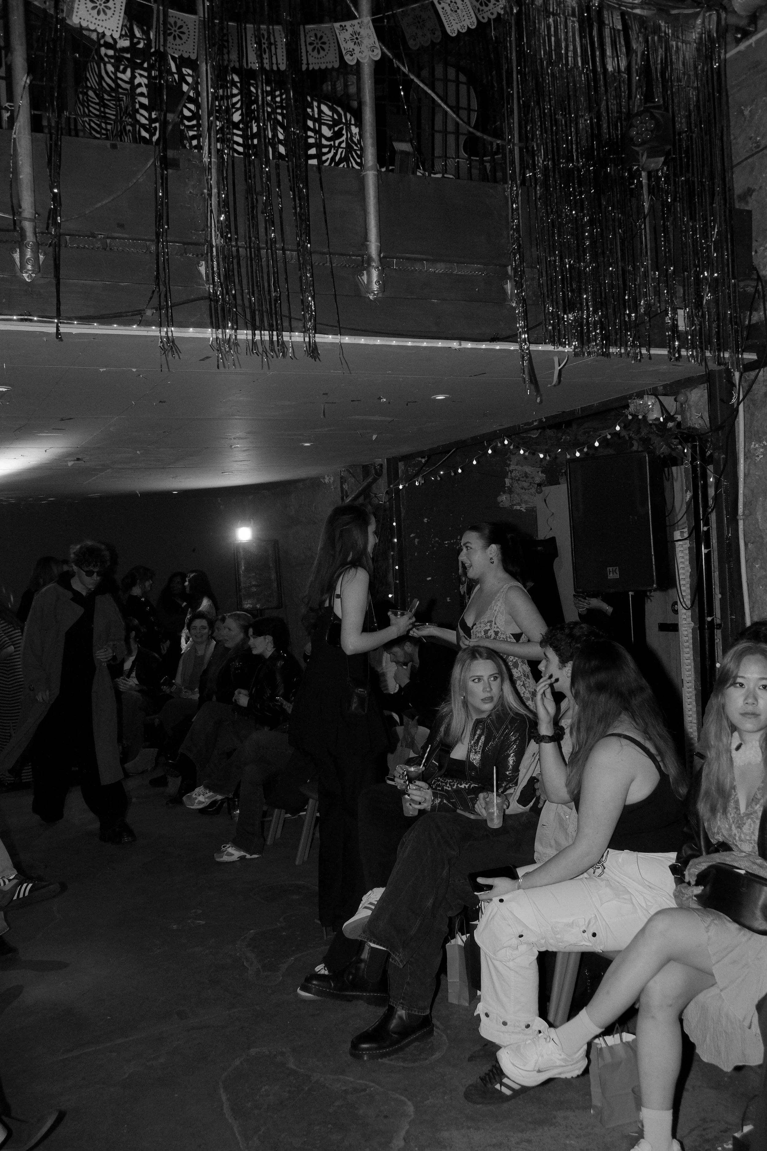People sitting and standing at a party or social gathering in an indoor venue with decorative hanging streamers and string lights.
