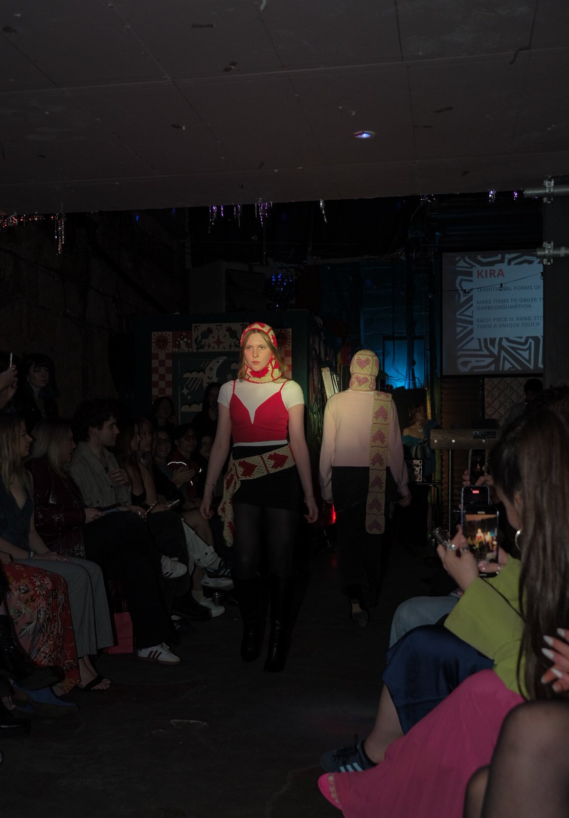 Models walk on runway during a fashion show, surrounded by seated audience.