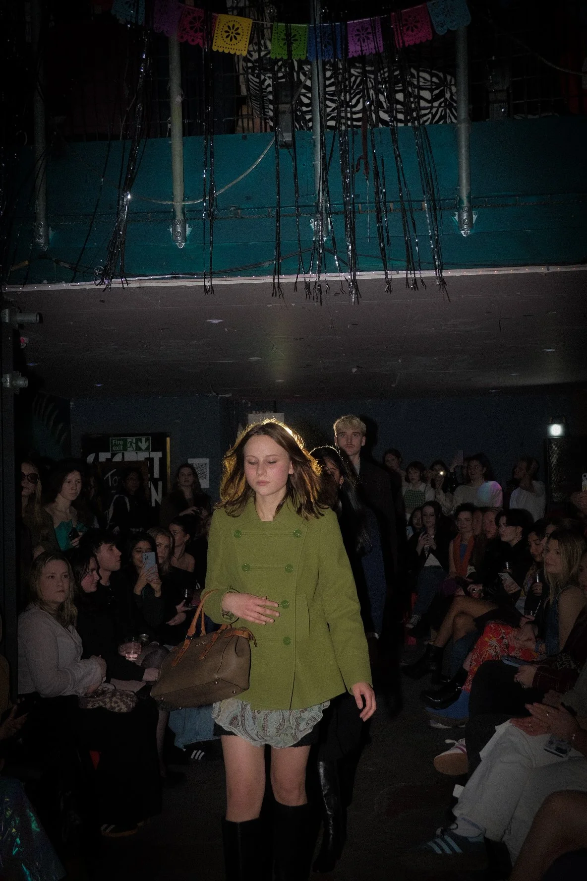 A young woman with wavy hair, wearing a green blazer, a short skirt, and black boots, walking in front of an audience at a fashion show, with people seated on both sides and some taking pictures.