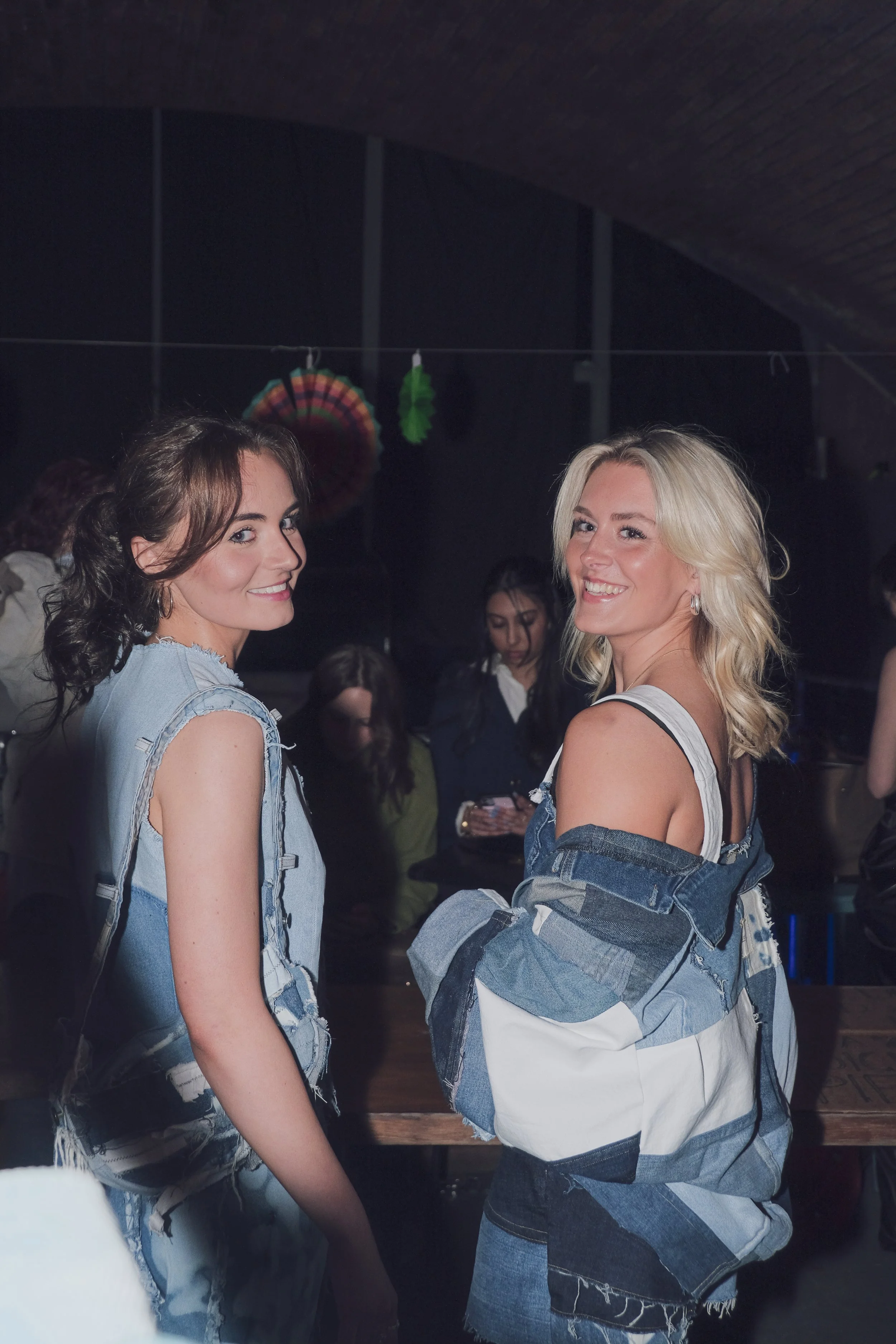 Two women smiling and posing for the camera at a social gathering or party, with other people in the background.