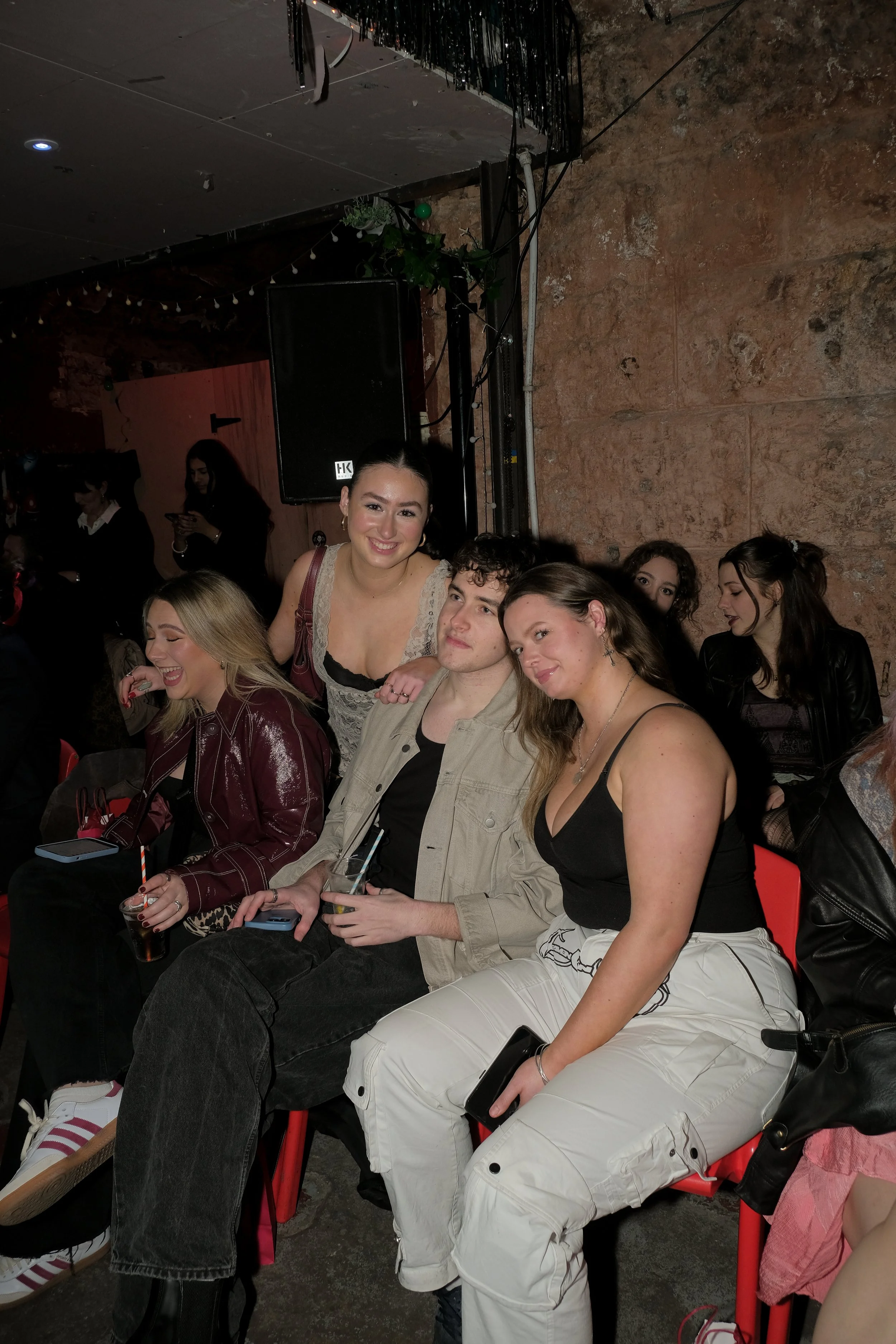 Group of young people sitting on red chairs at a social event, with a stone wall and a speaker in the background.