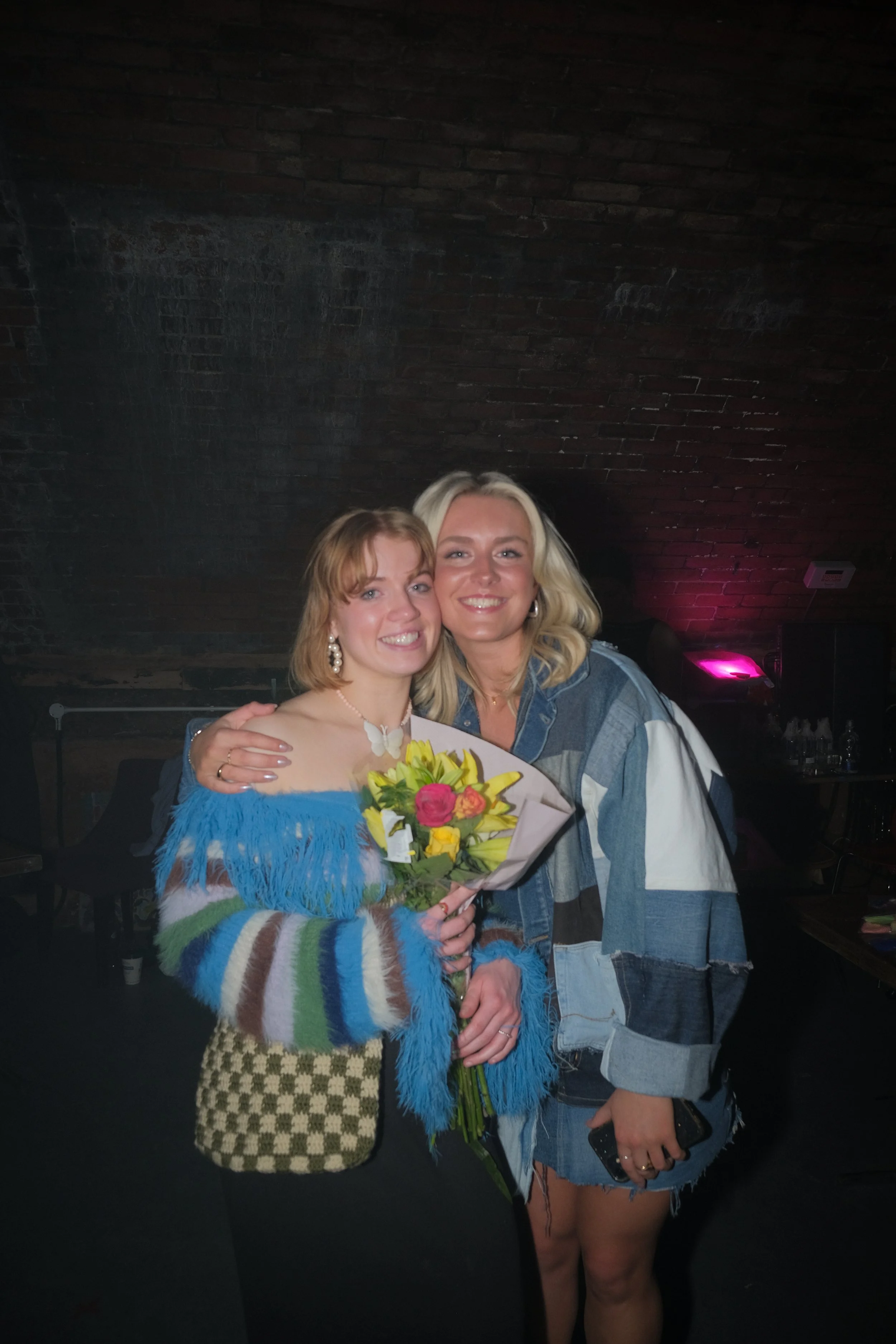Two women smiling and hugging, one holding a bouquet of colorful flowers, in a dimly lit indoor setting with a brick wall background.