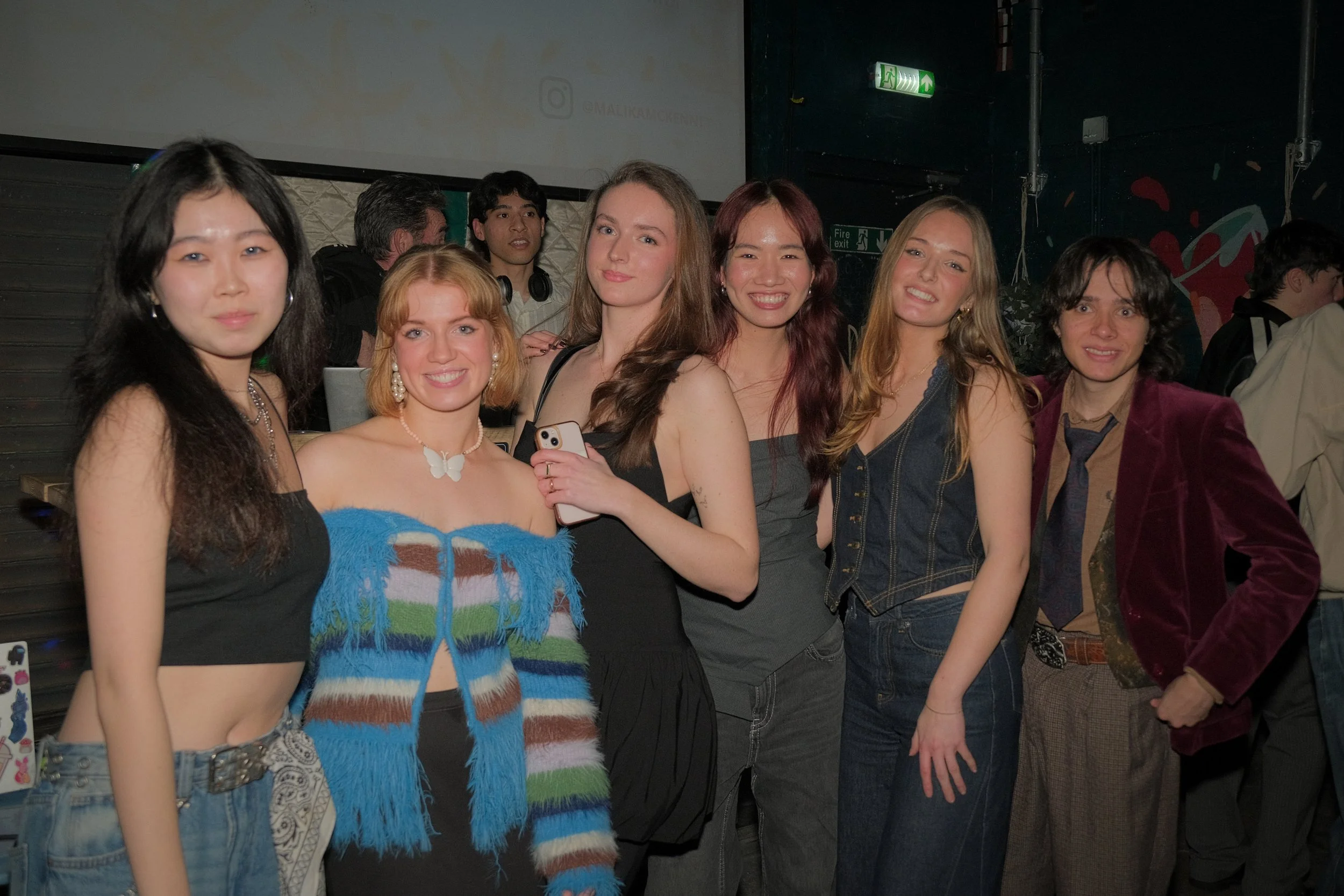 Seven young women posing together at a party or event, smiling, with a DJ and other people in the background.