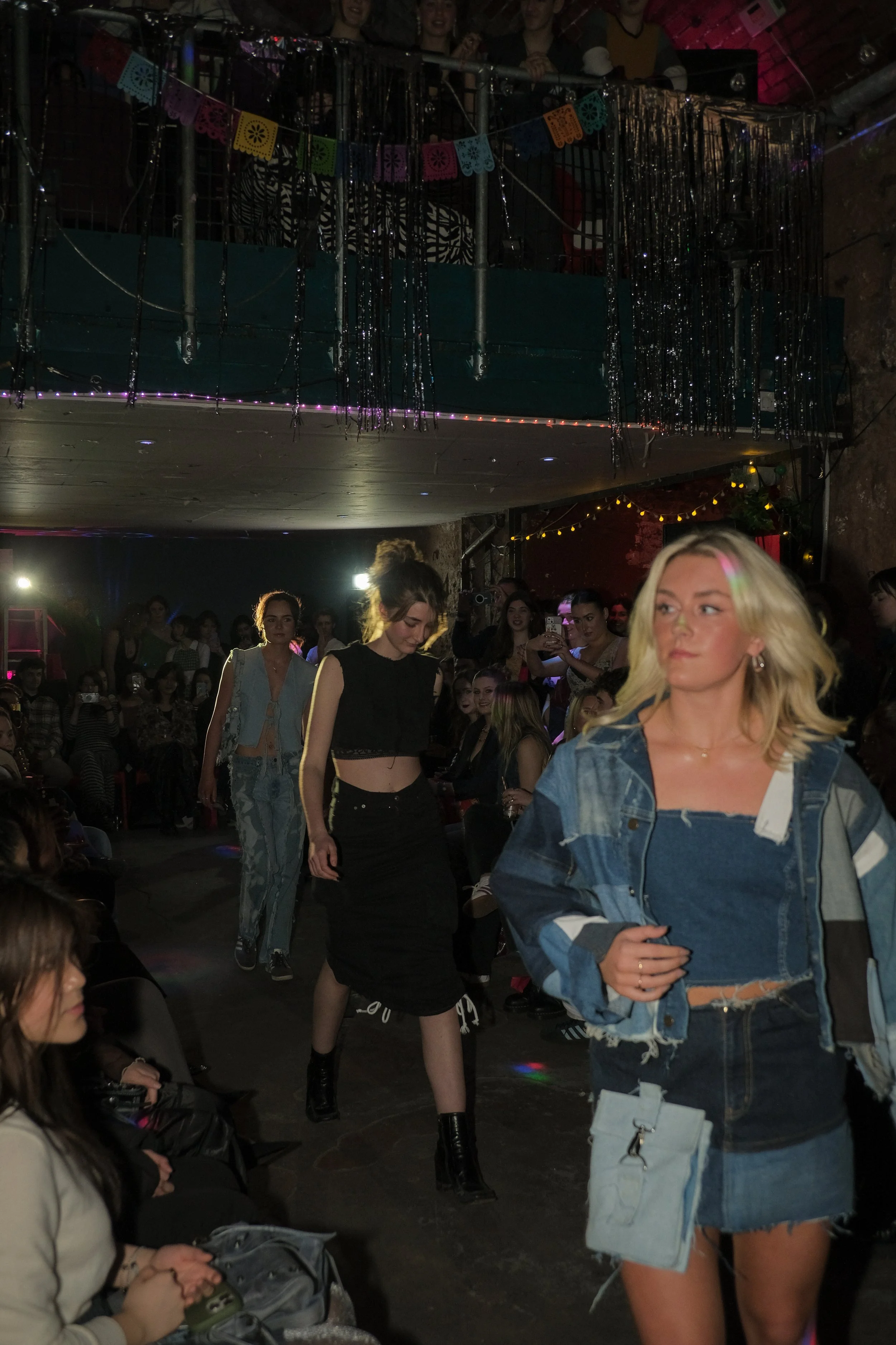 Fashion models walking on a runway during a fashion show with an audience seated on either side, colorful bunting hanging from the ceiling, and decorative lighting.