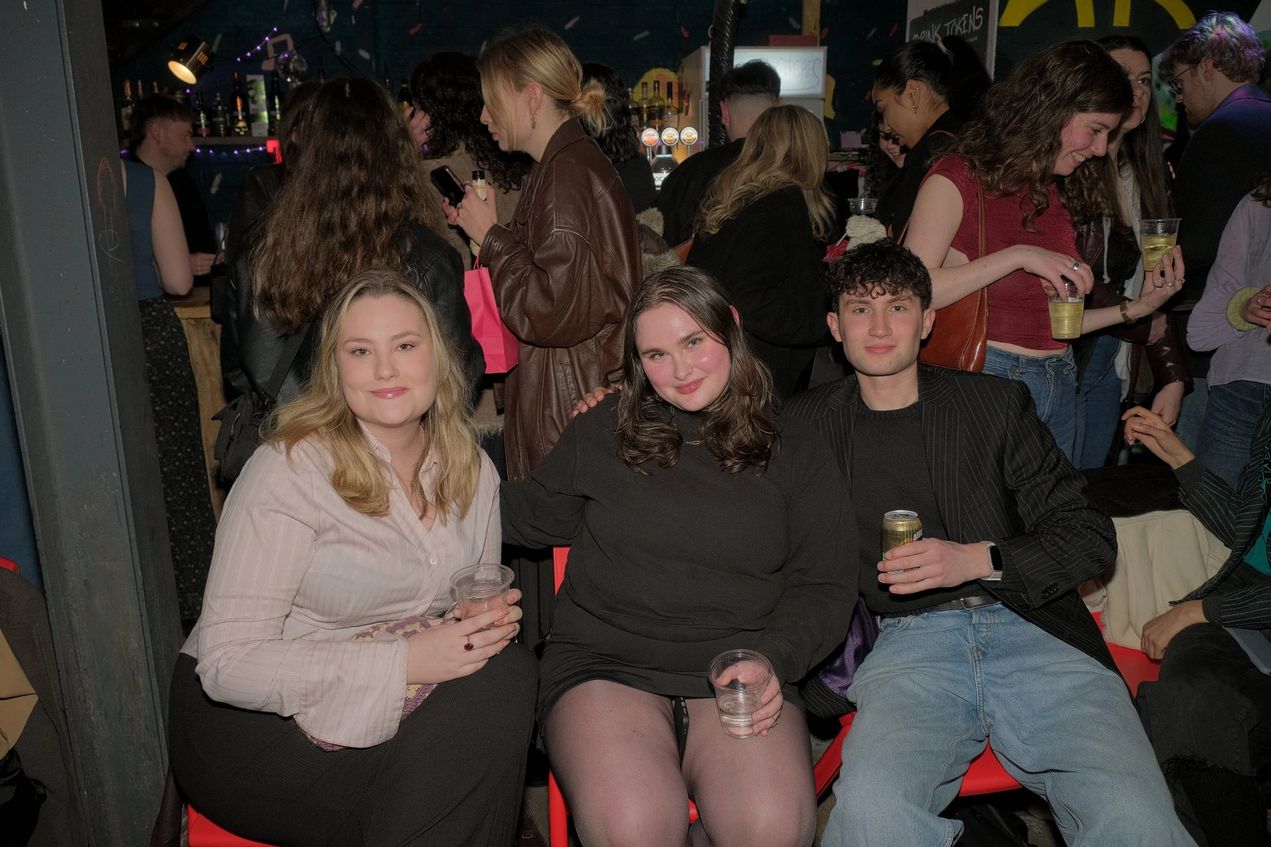 Three young adults sitting together at a crowded bar or club, two women and one man, smiling and holding drinks, with a lively background of other patrons and a bar area.