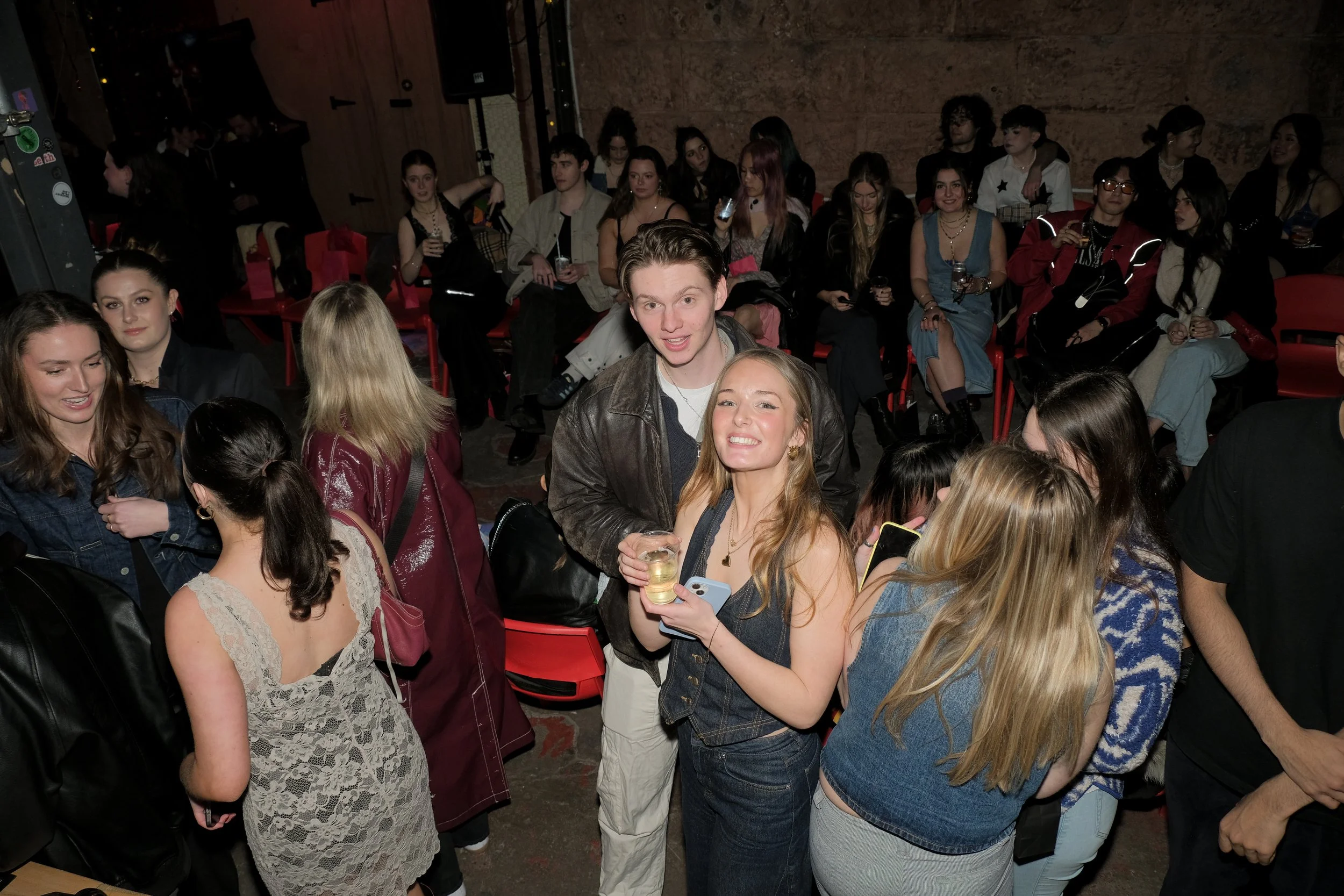 Group of young people at a social gathering, some holding drinks, with a crowd sitting in the background.