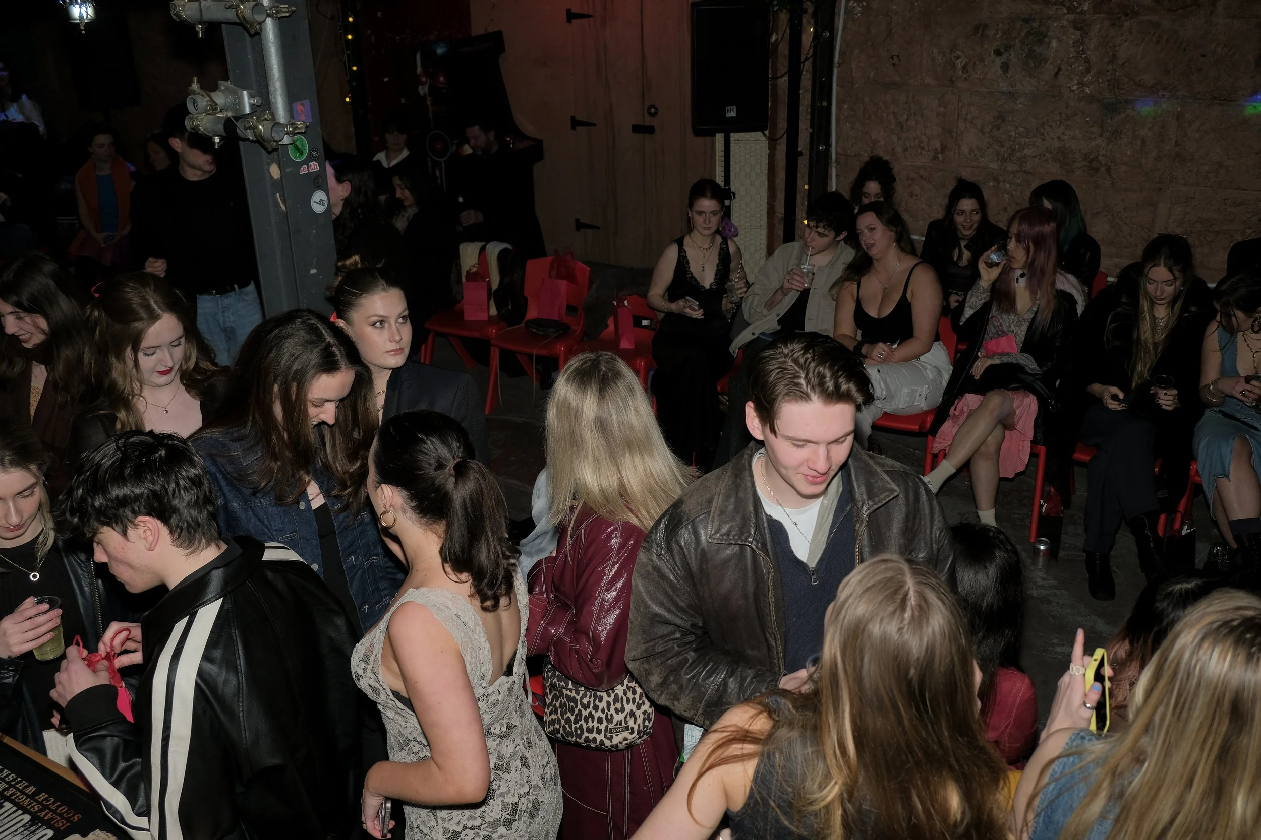 A crowded indoor event with many young people socializing and sitting on red chairs. Some people are looking at their phones, and there is a mix of casual and slightly formal attire.