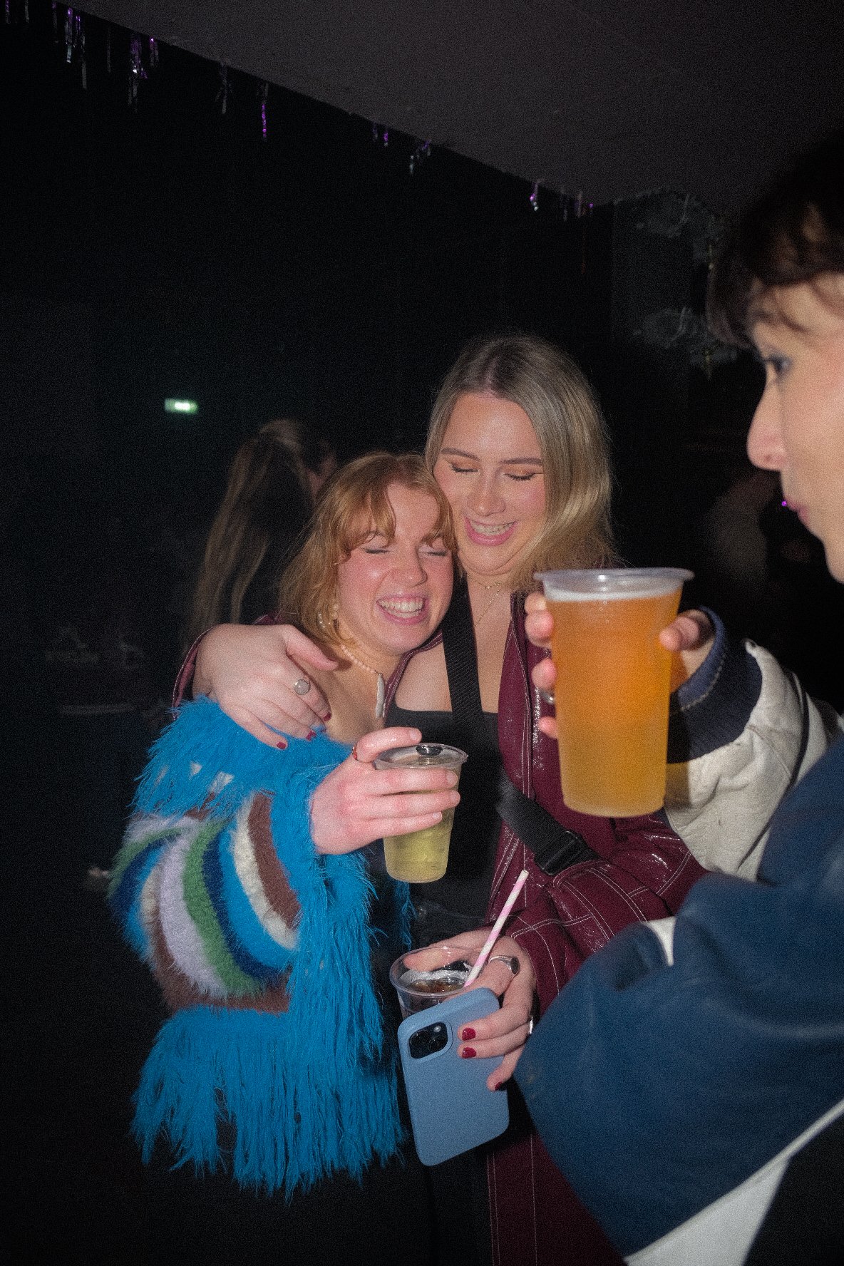 Two women smiling and hugging at a party, holding drinks, with a man on the right holding a cup of beer, in a dark room with purple string lights.