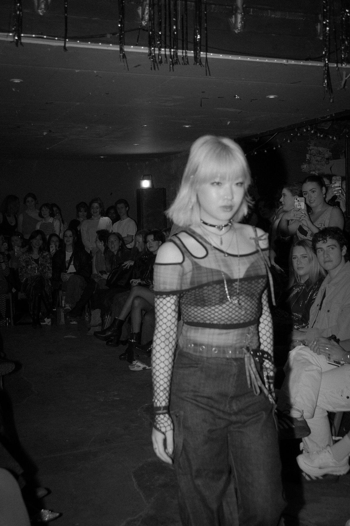 A woman with blonde hair walking down a runway during a fashion show, with an audience of mostly young women and a few men watching in the background, some taking photos.