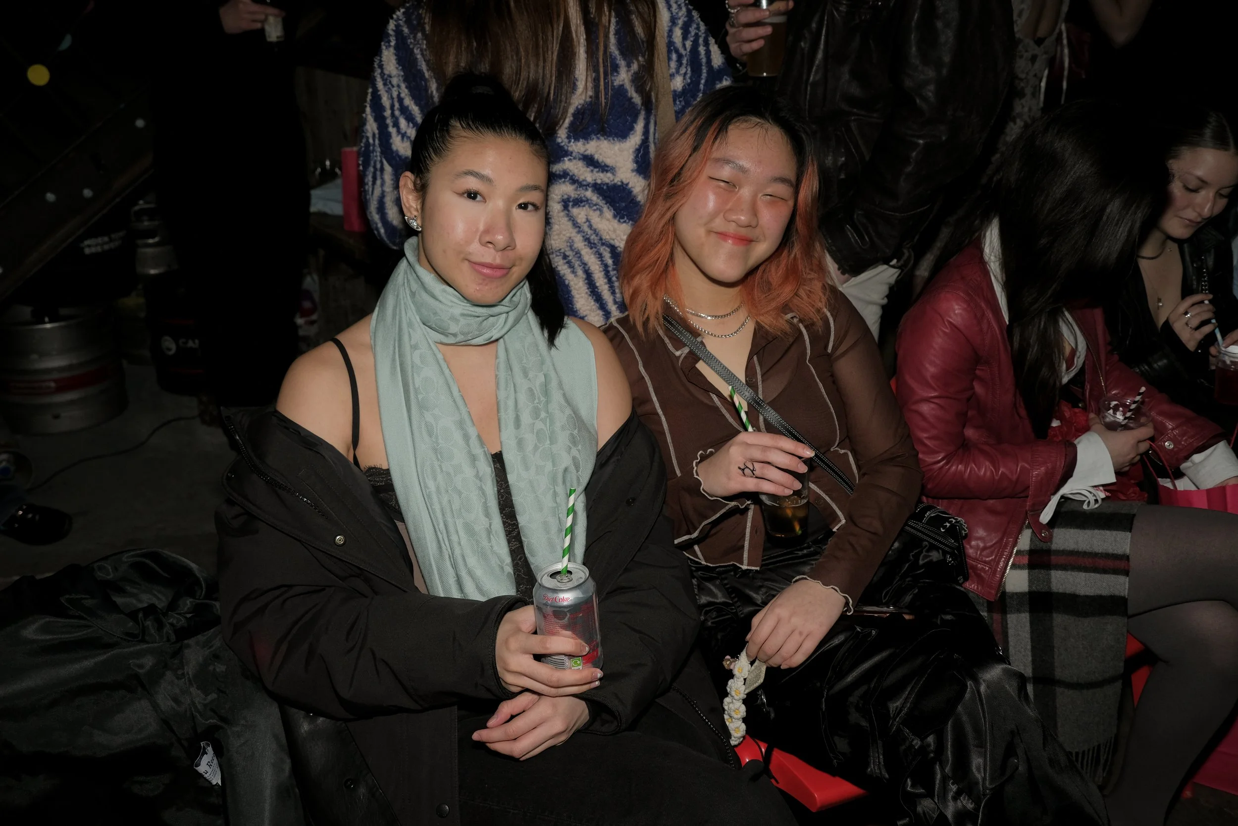 Two young women sitting on a bench at a social event, holding cans with straws, surrounded by other people.