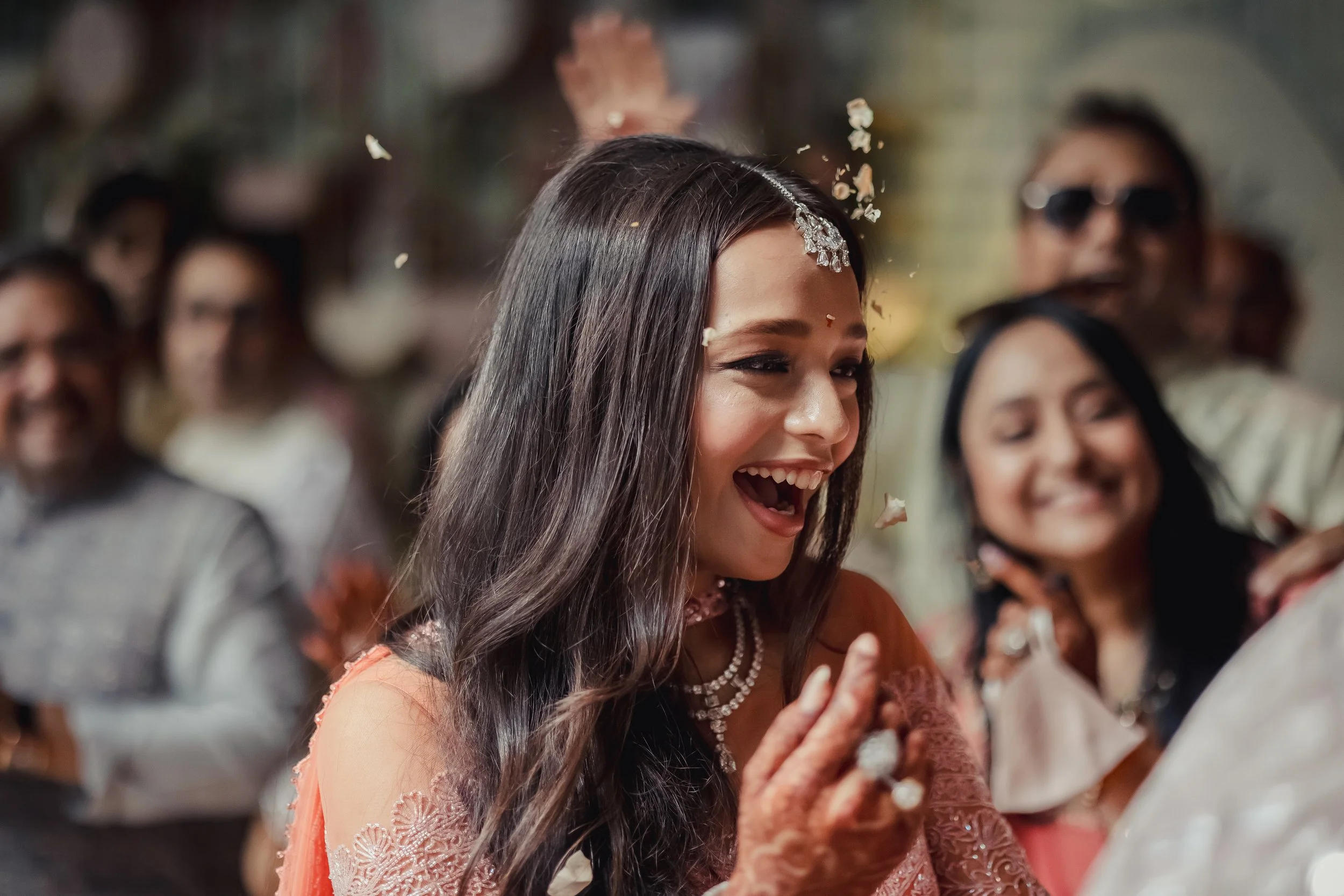 A woman with long dark hair, wearing traditional attire and jewelry, smiling and surrounded by celebrating people at a festive event.