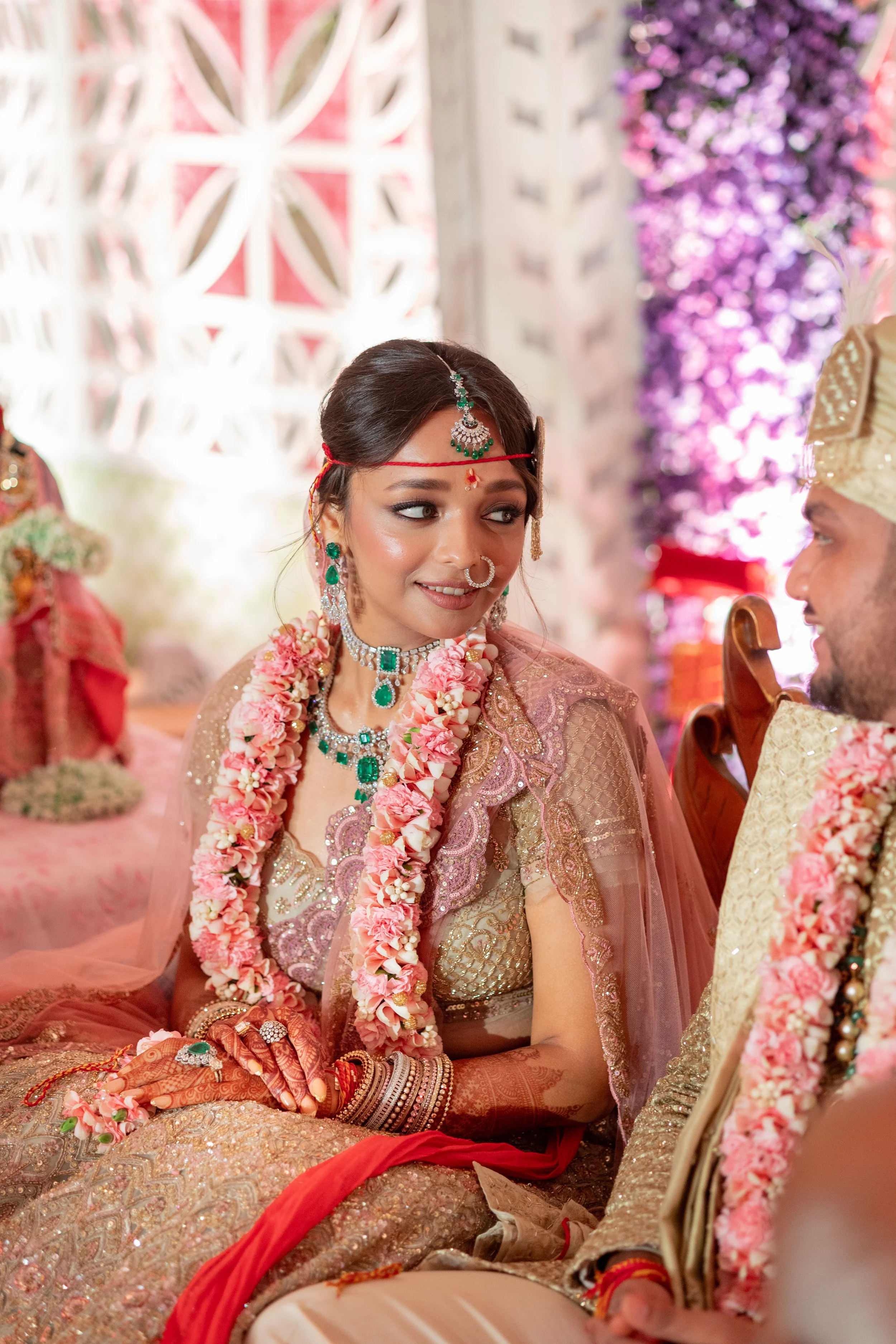 A bride and groom dressed in traditional Indian wedding attire, sitting during their wedding ceremony. The bride wears elaborate jewelry and garlands of flowers, while the groom is in a decorated outfit with a turban. They are exchanging looks amidst