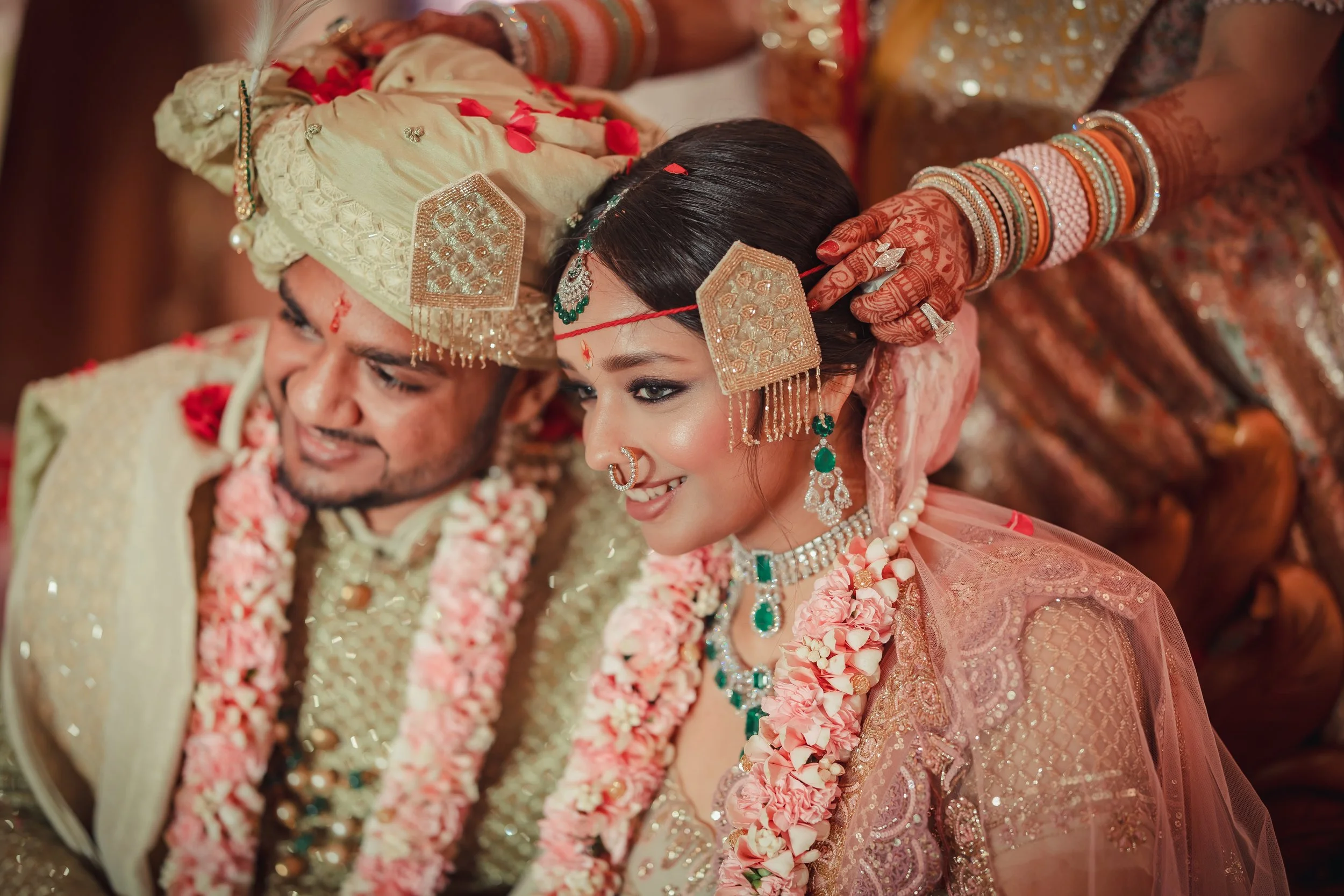 An Indian bride and groom dressed in traditional wedding attire, sitting closely together during their wedding ceremony. The bride wears an ornate lehenga with jewelry, and the groom wears a sherwani and turban, both decorated with flower garlands.