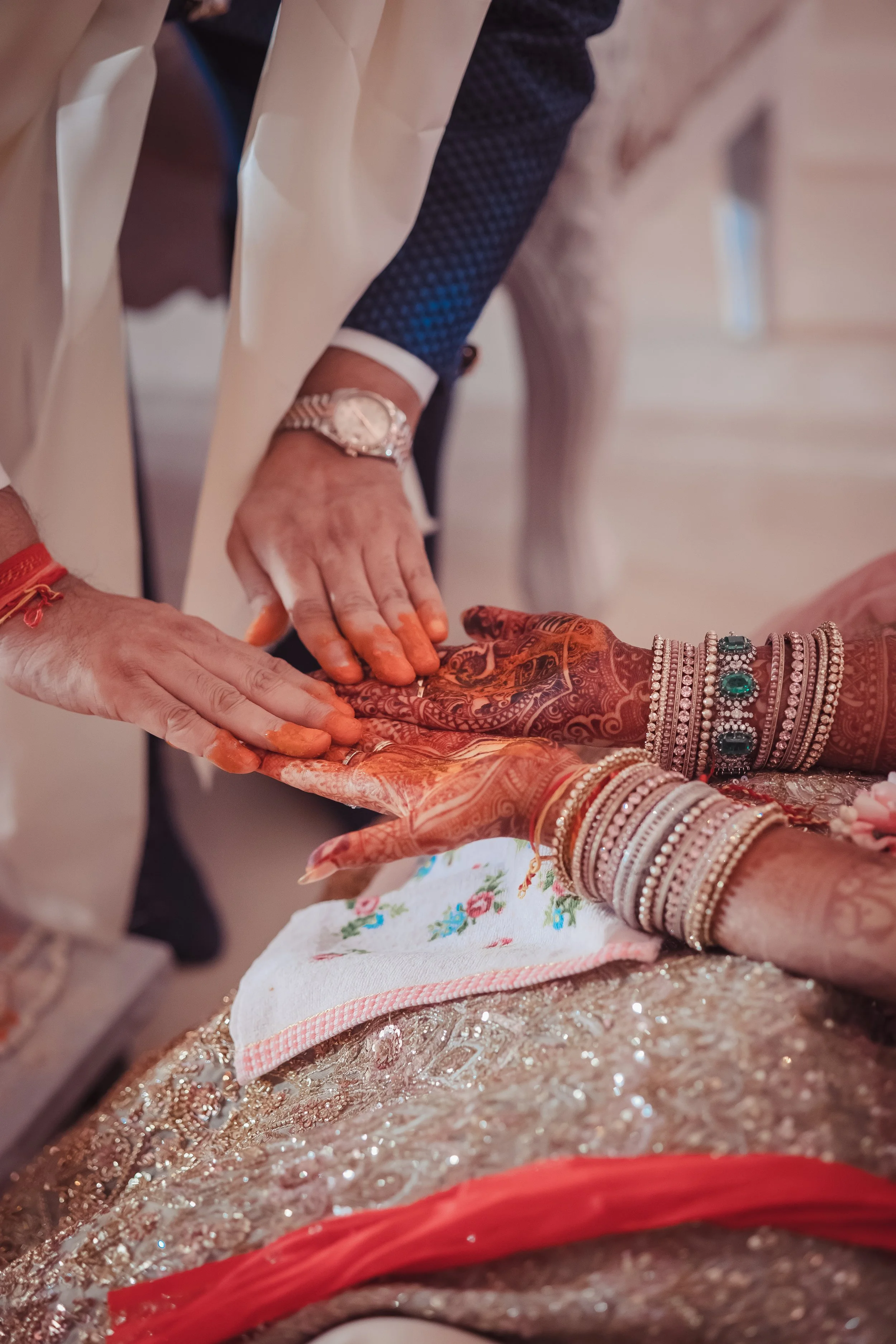 Close-up of a traditional Indian wedding ceremony showing a bride with decorated hands and jewelry, and a person, possibly the groom, touching her hands with the couple's hands resting on a cloth-covered surface.