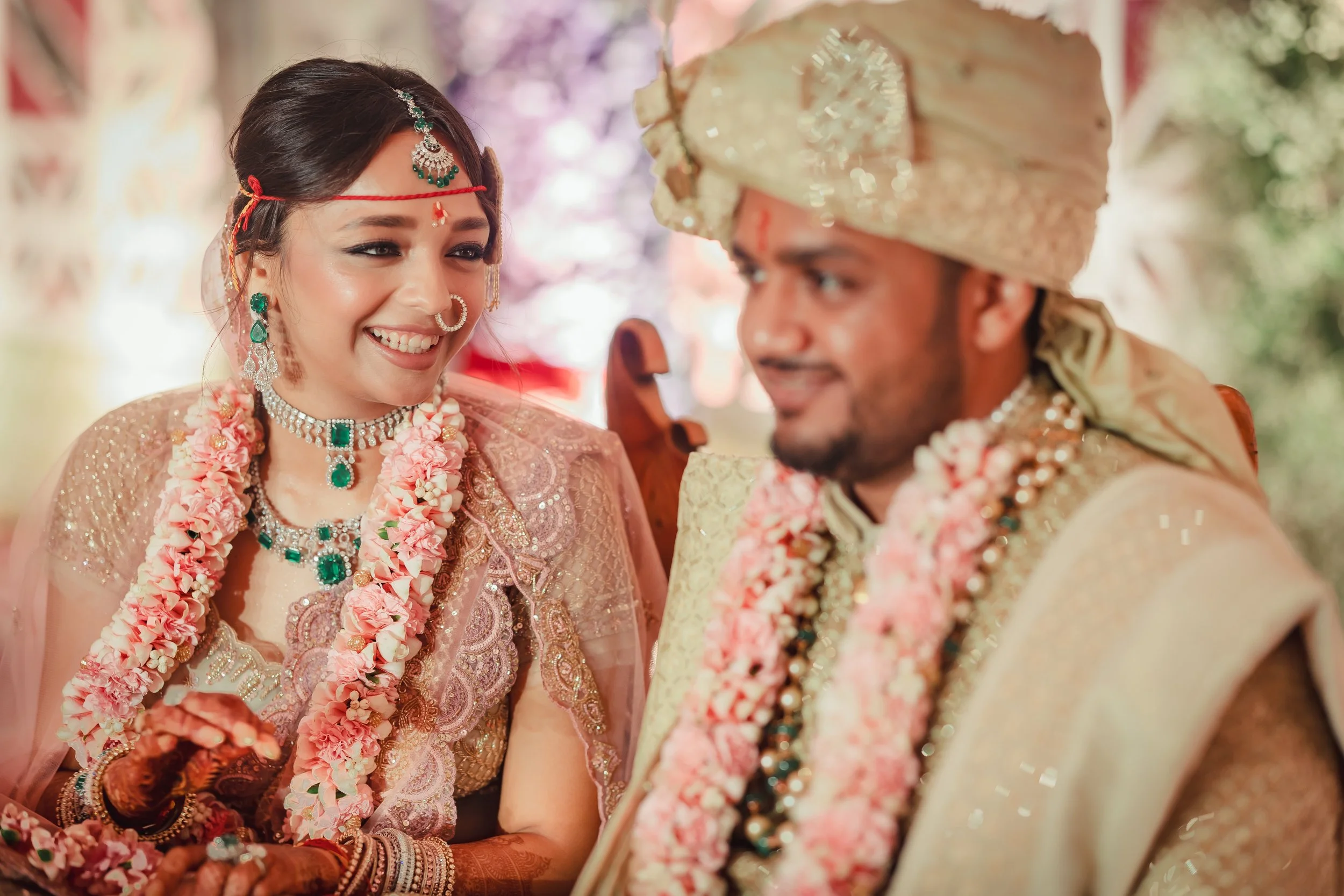Indian bride and groom in traditional wedding attire, sitting together, smiling, with floral garlands around their necks.