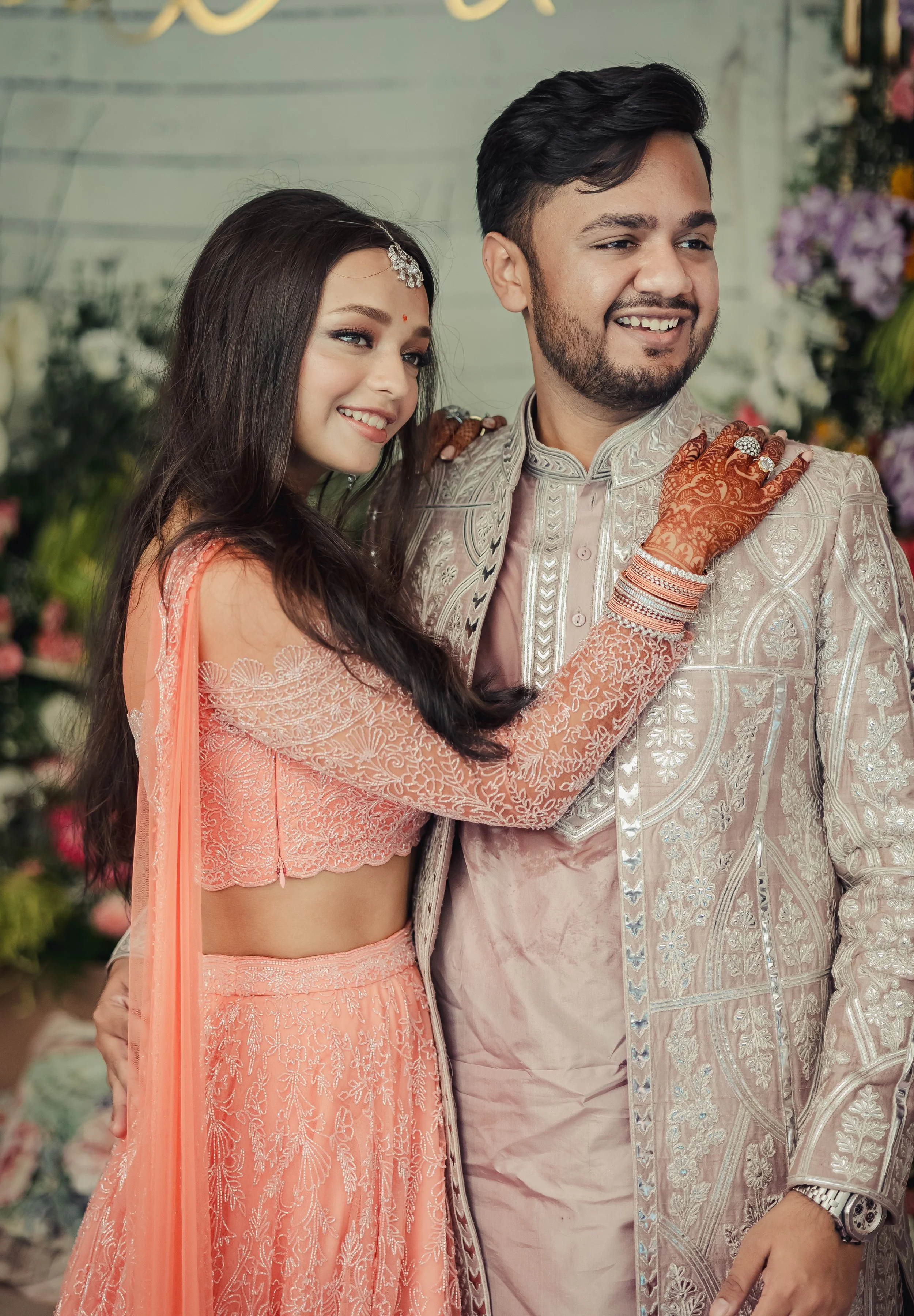 A happy couple dressed in traditional Indian wedding attire, with the woman in peach embroidery and the man in a silver embroidered sherwani, smiling and posing for a photo at their wedding.