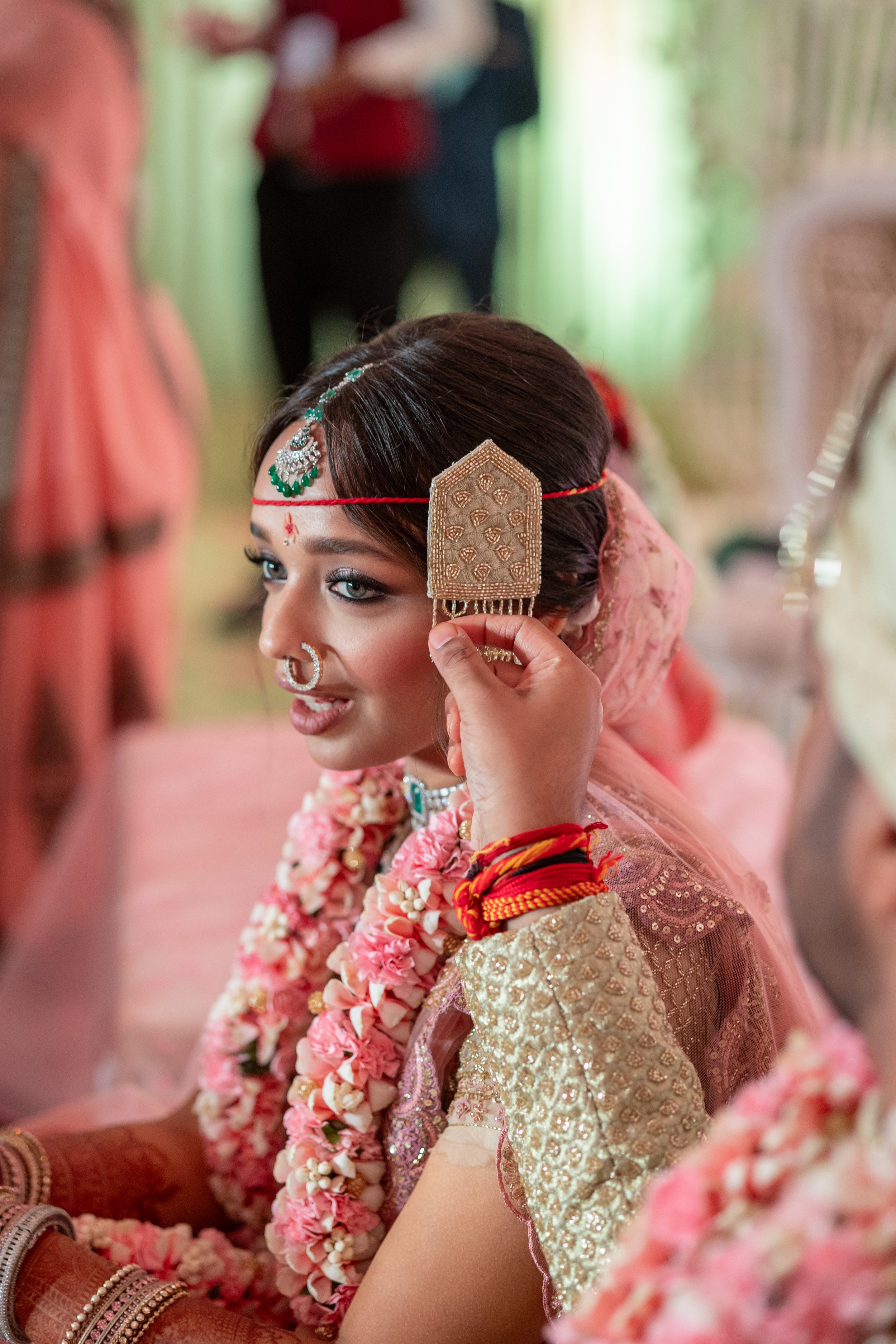 A bride with earrings, nose ring, and decorative headpiece during a traditional wedding ceremony, with a person attaching an ornamental piece to her hair.