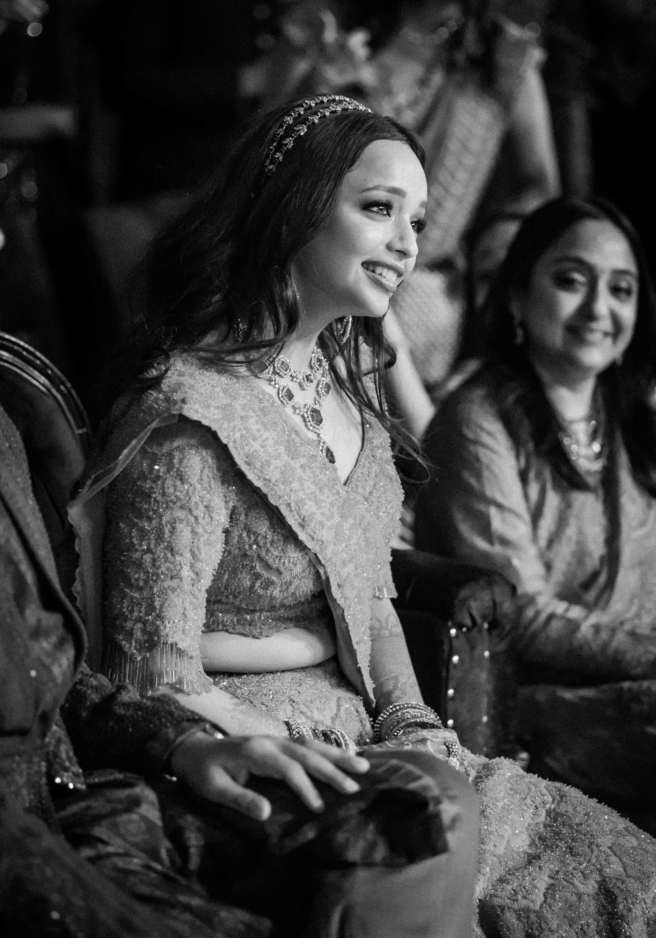 A woman dressed in traditional Indian attire, wearing jewelry, smiling and seated next to another woman, at an event.