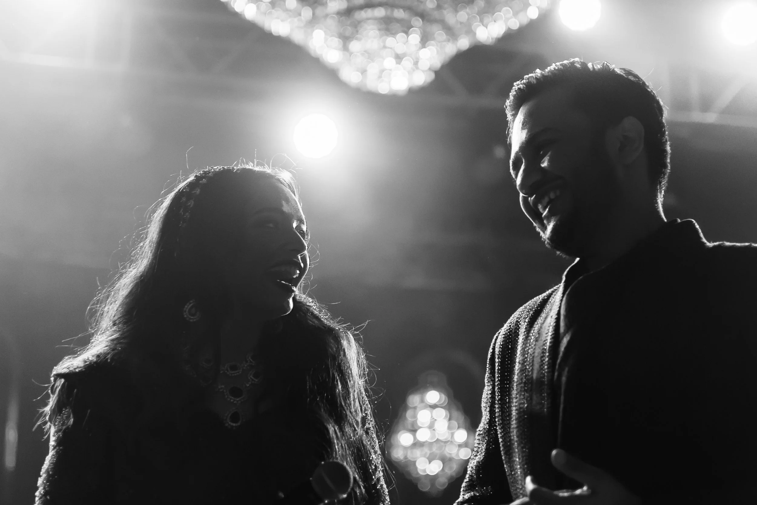 Black and white photo of a man and a woman smiling at each other on a stage with bright lights overhead.