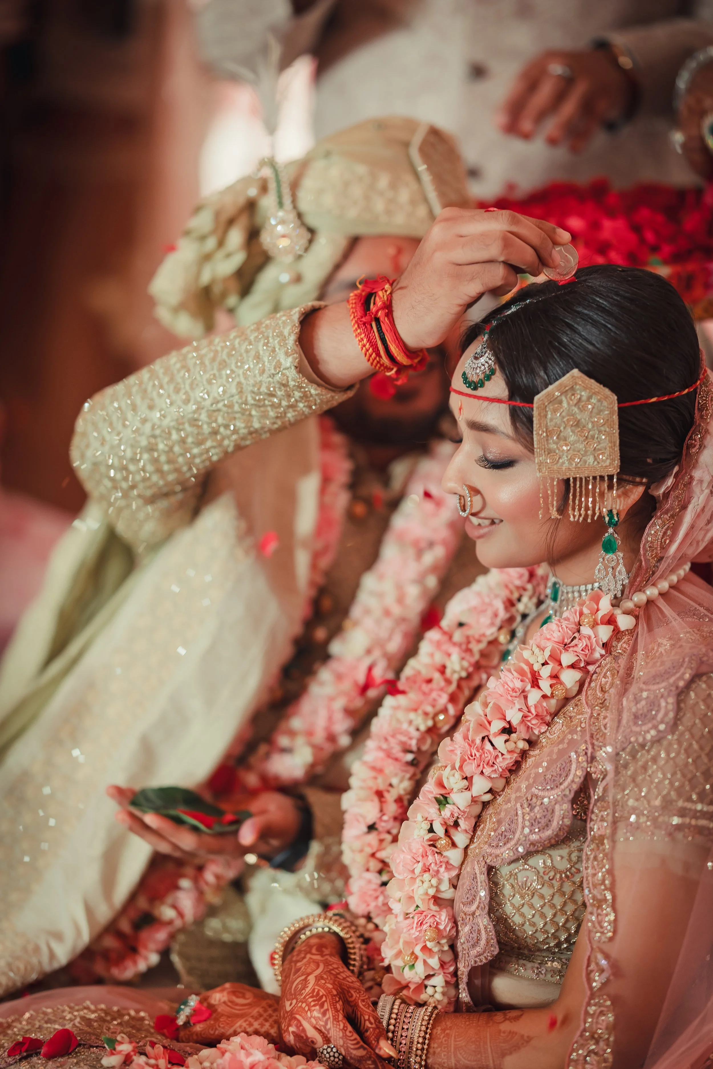 A bride with traditional Indian attire and jewelry, smiling during her wedding ceremony as a person adjusts her headpiece.