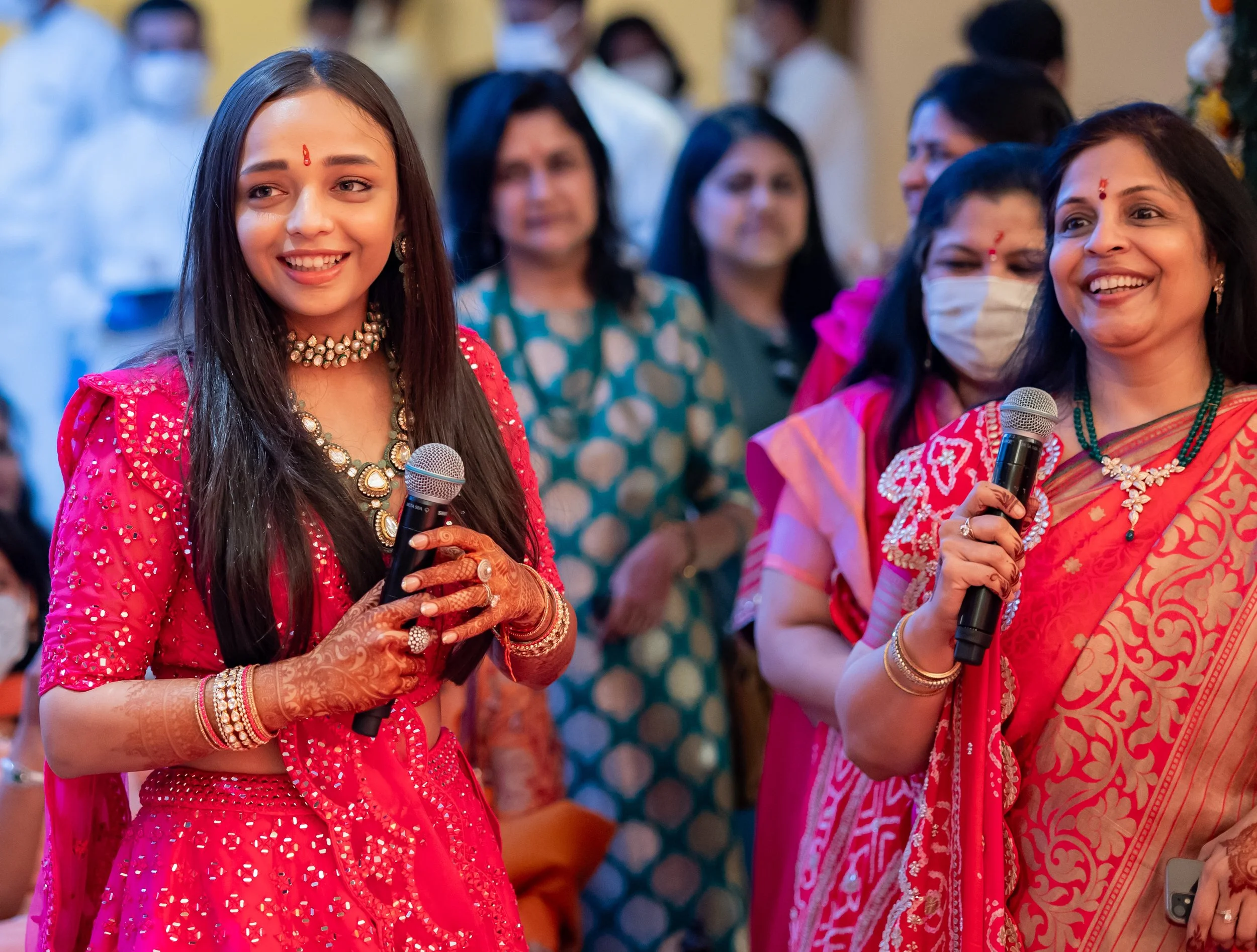 Young woman in a bright pink traditional dress and jewelry holding a microphone, surrounded by women in colorful traditional attire, at a celebration or cultural event.