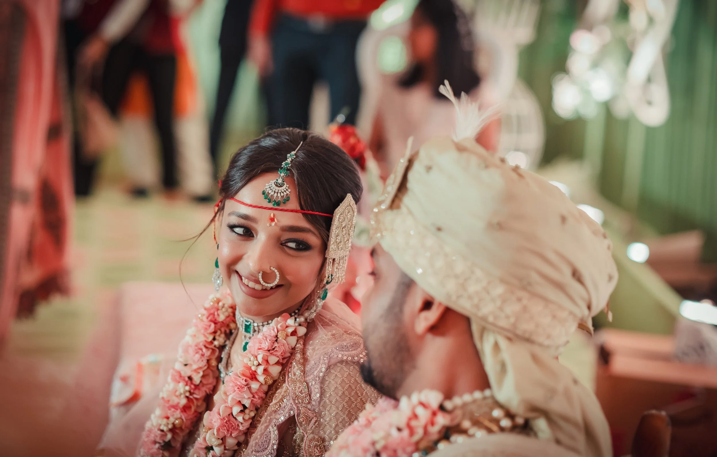 A bride and groom at a traditional Indian wedding, dressed in elaborate attire and jewelry, smiling at each other with the bride wearing a floral garland.