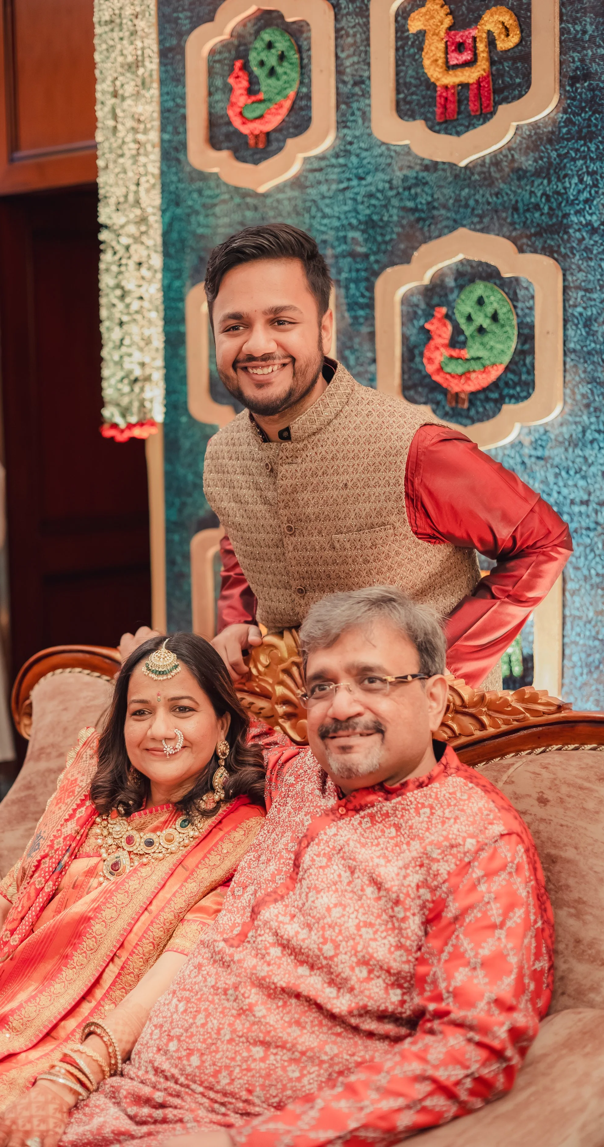 A family celebrating a traditional Indian wedding, with a woman in a saree and jewelry, a man in a patterned kurta, and a young man in a vest, all smiling and sitting on a decorated sofa with festive wall art in the background.