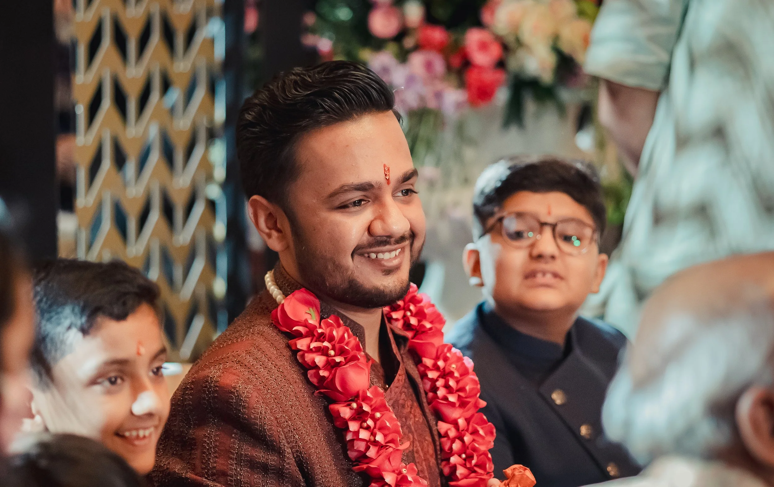 A group of people dressed in traditional Indian attire at a celebration, with a man in the center wearing a maroon sherwani and a red flower garland, smiling.