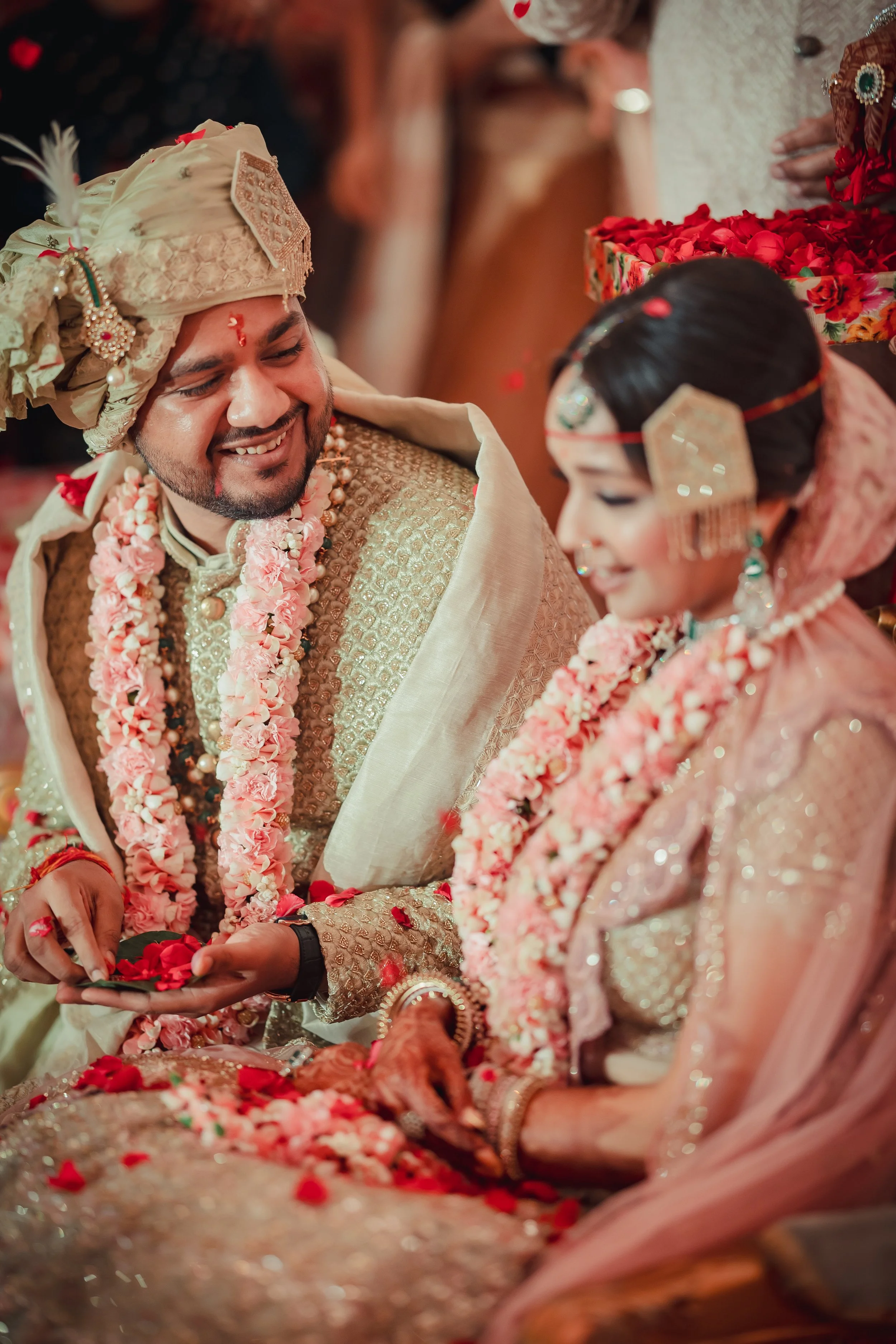 Indian wedding ceremony with bride and groom dressed in traditional attire, exchanging flower petals and garlands, surrounded by guests.