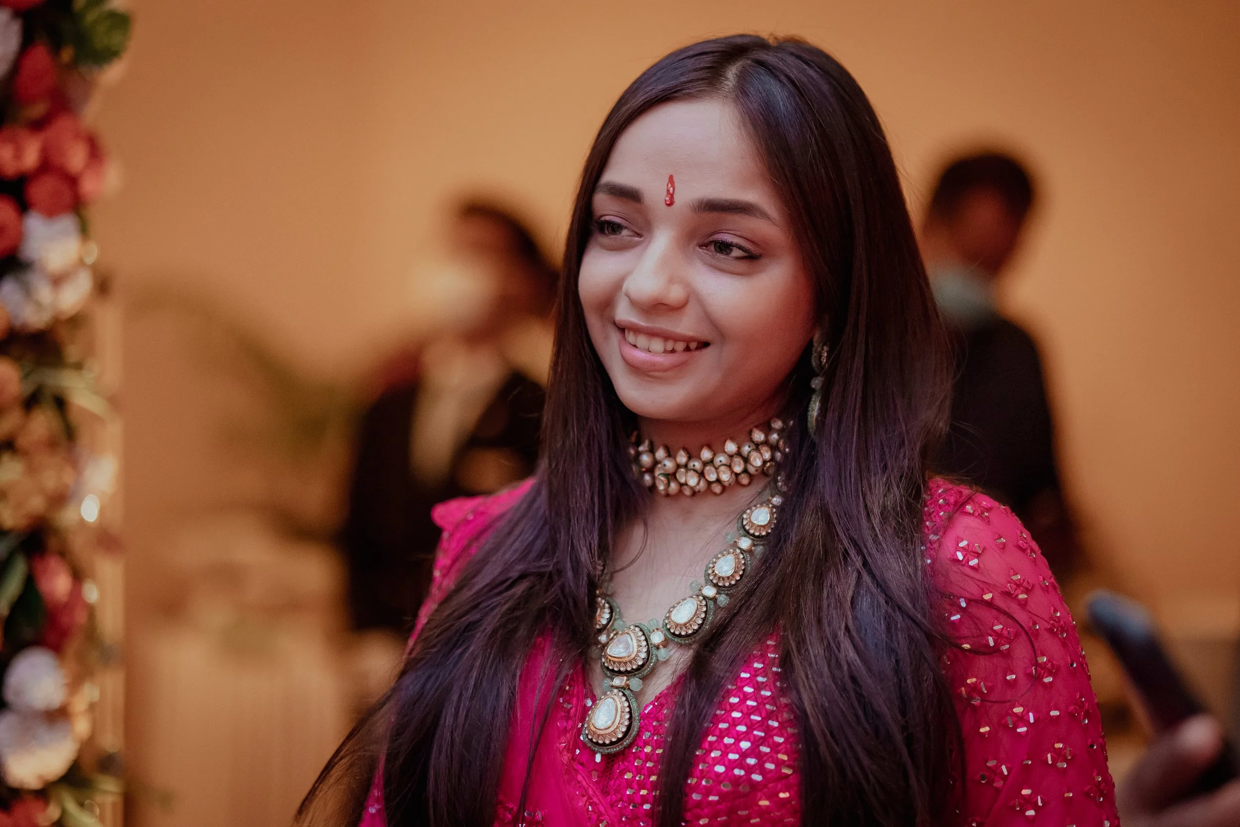 A young woman with long dark hair and light skin, dressed in a bright pink traditional outfit embellished with gold accents, wearing a pearl and gold necklace and earrings, and a small red bindi on her forehead, smiling at a festive event.