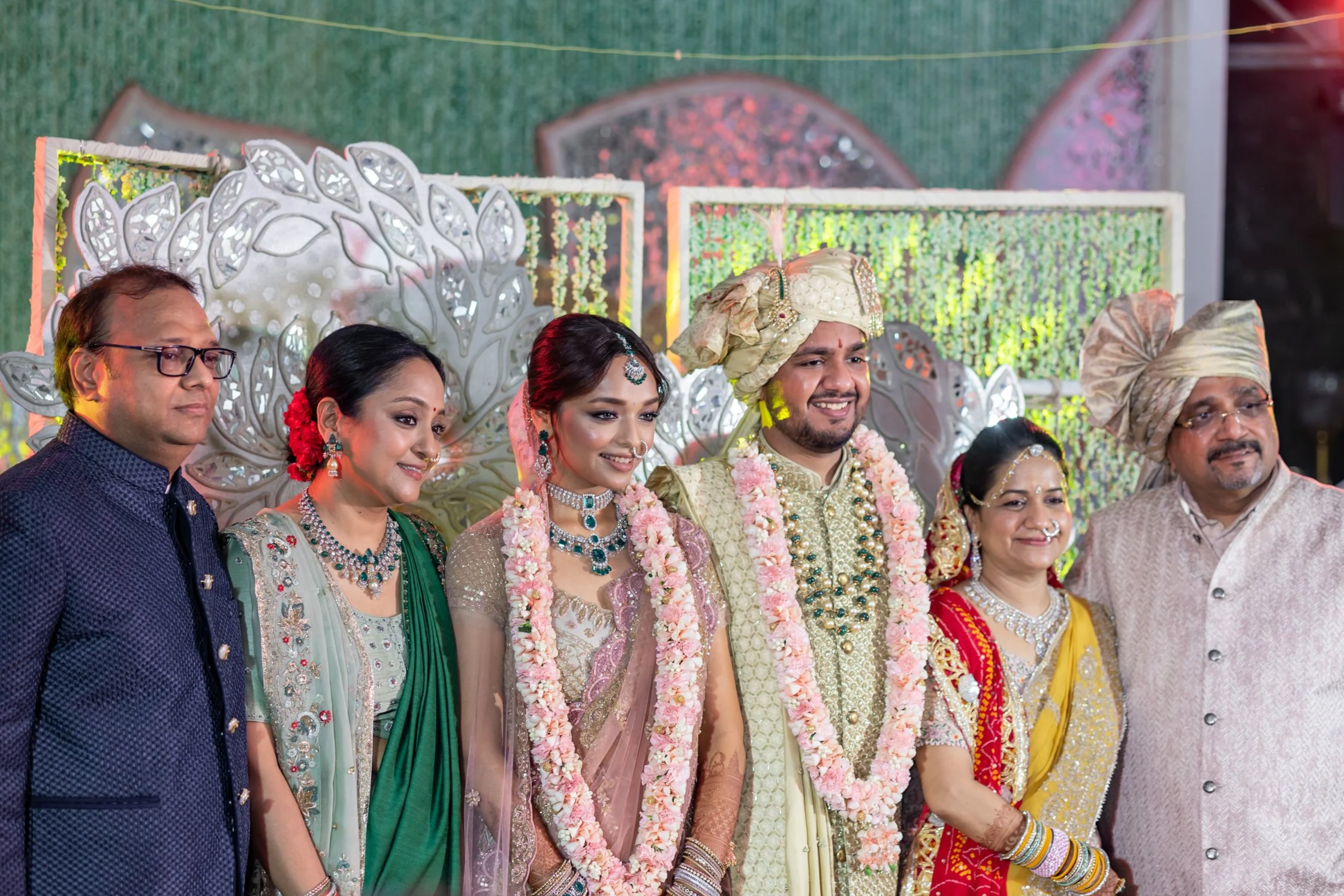 A group of seven people dressed in traditional Indian wedding attire, standing in front of decorative backdrop, smiling for the photo.
