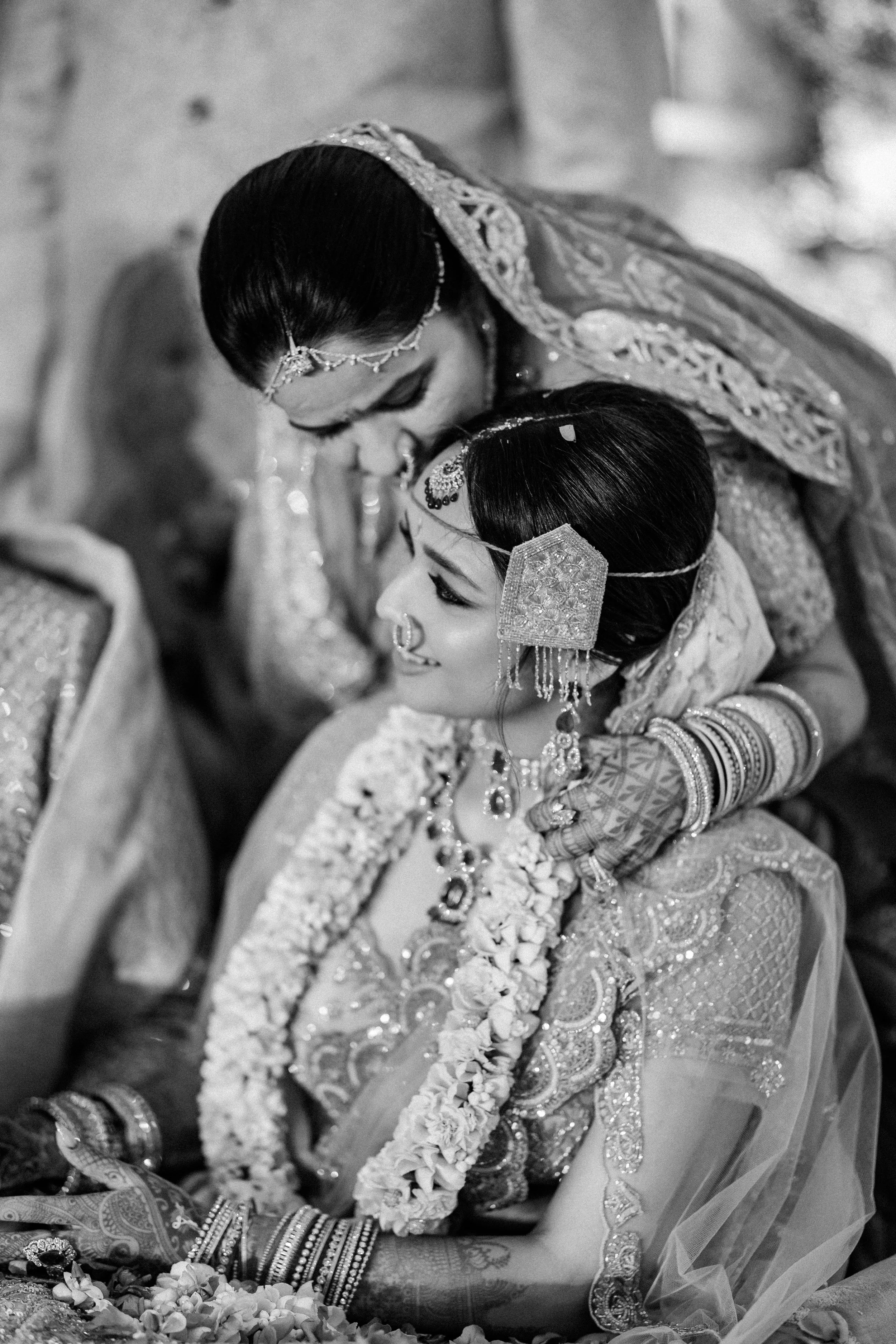Two women dressed in traditional Indian wedding attire, engaged in a tender moment, with one woman leaning over the other.