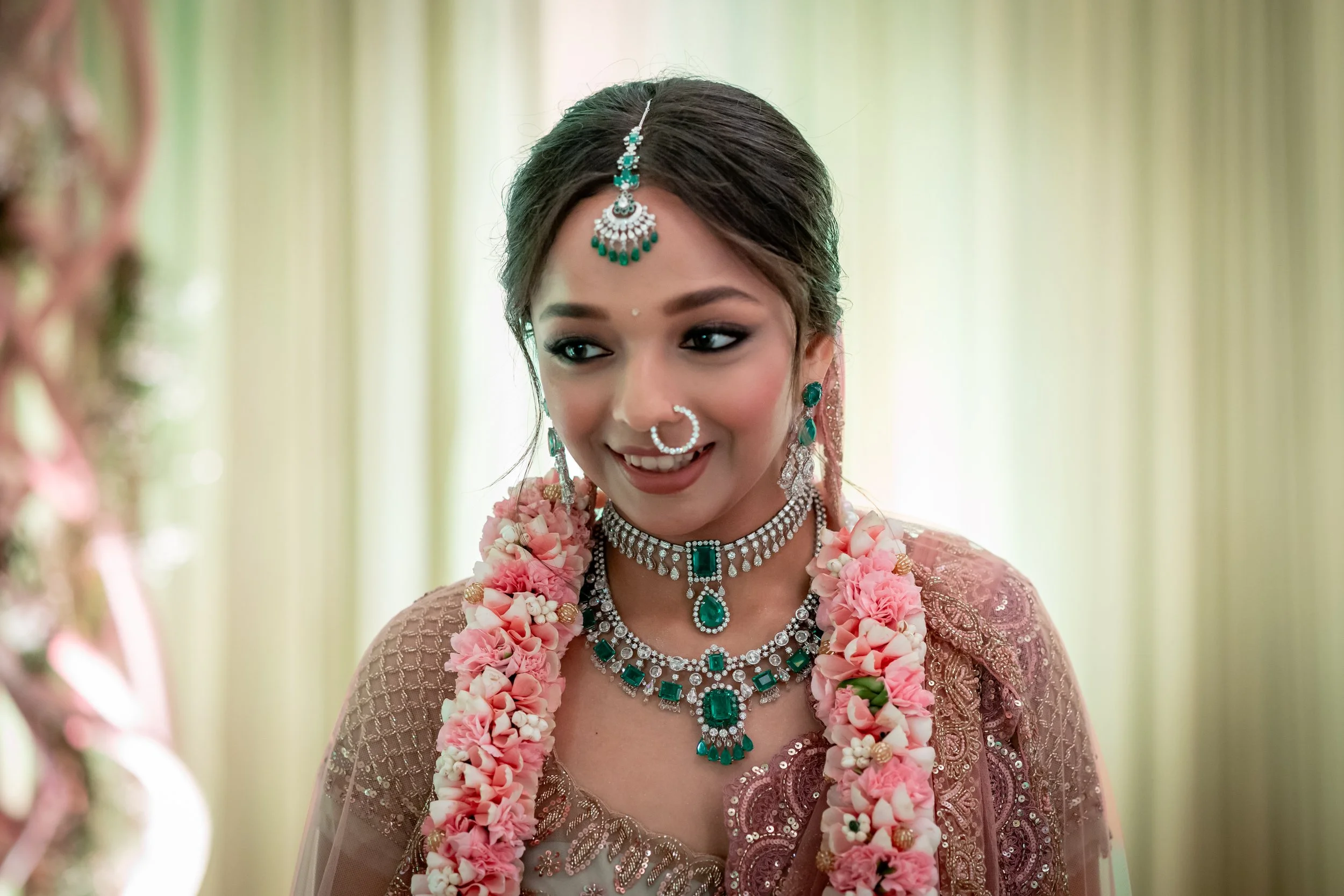 A young woman dressed in traditional Indian attire, wearing multiple necklaces, earrings, a nose ring, and a forehead ornament, with flower garlands around her neck, smiling during a celebratory event.