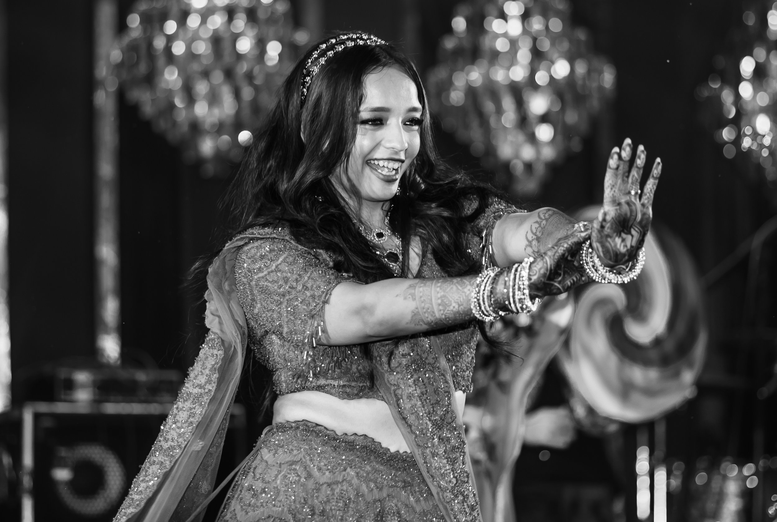 A woman with long dark hair wearing a heavily embellished traditional outfit, smiling and raising her hand in a greeting or wave, with intricate henna on her hand, jewelry on her wrists, and a blurred decorative background.