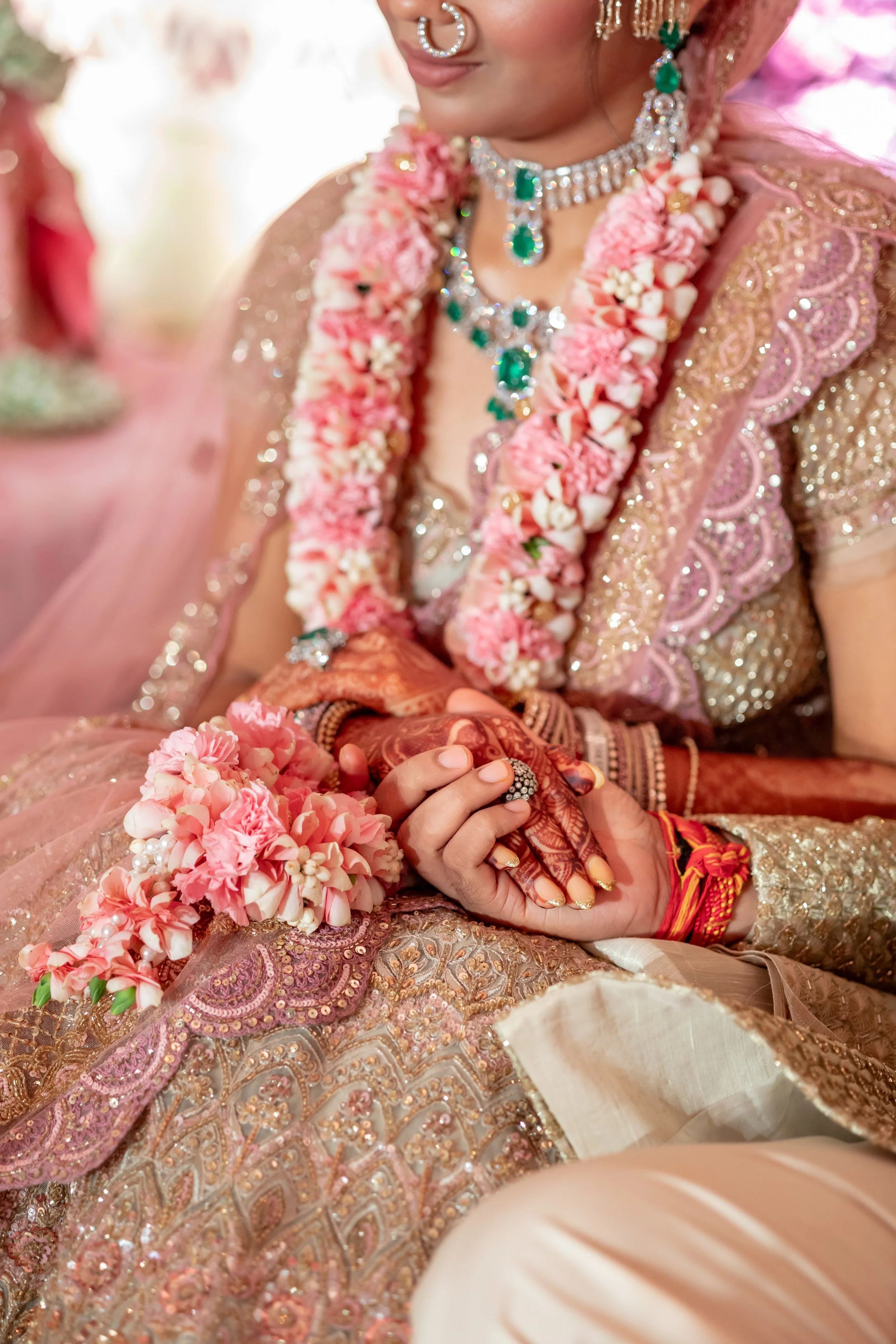 Close-up of a bride dressed in traditional Indian attire, adorned with jewelry, a floral garland, and intricate henna on her hands, during a wedding ceremony.