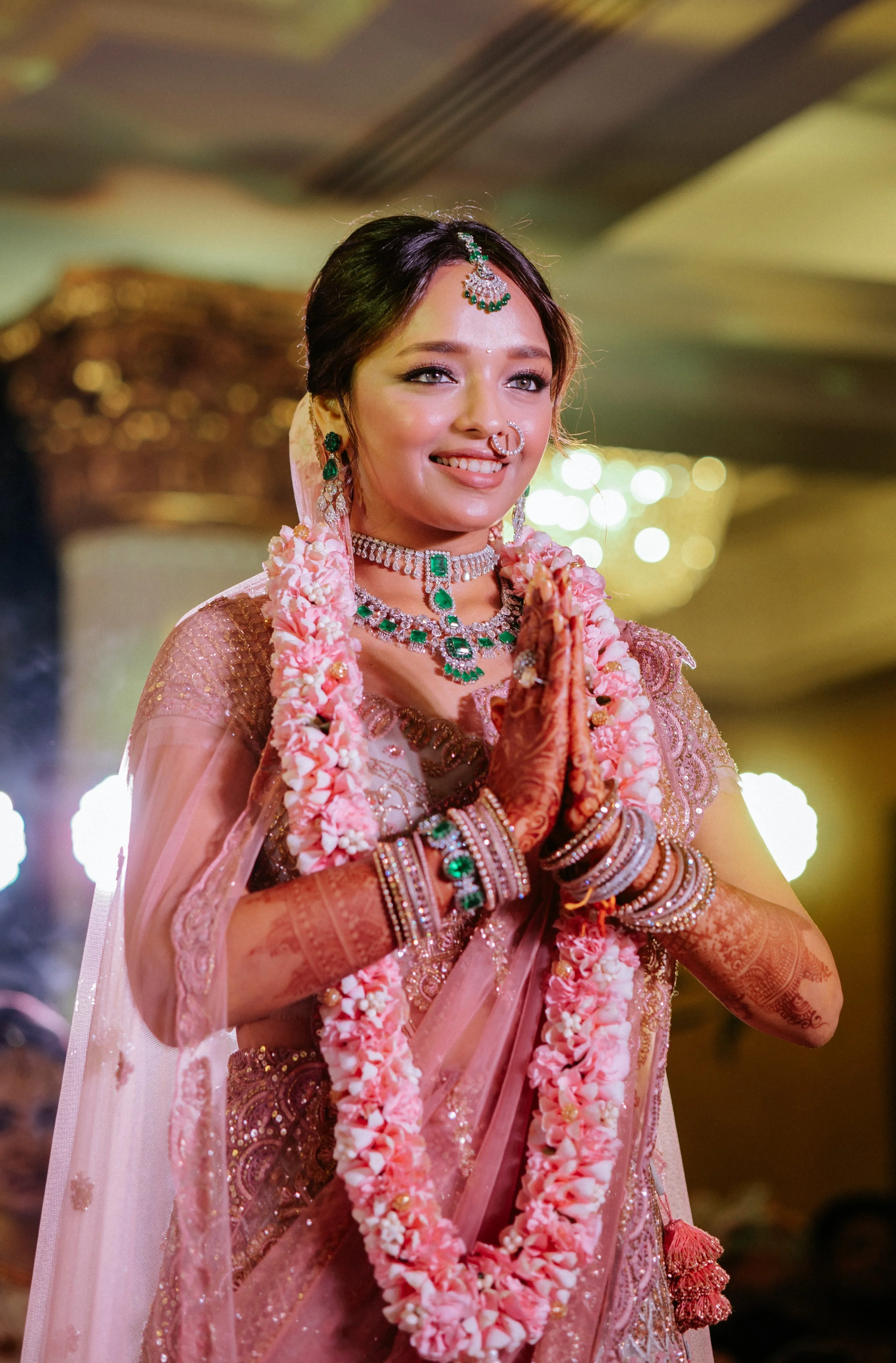 A bride dressed in traditional Indian wedding attire, smiling with hands in a prayer position, adorned with jewelry and flower garlands at an indoor celebration.