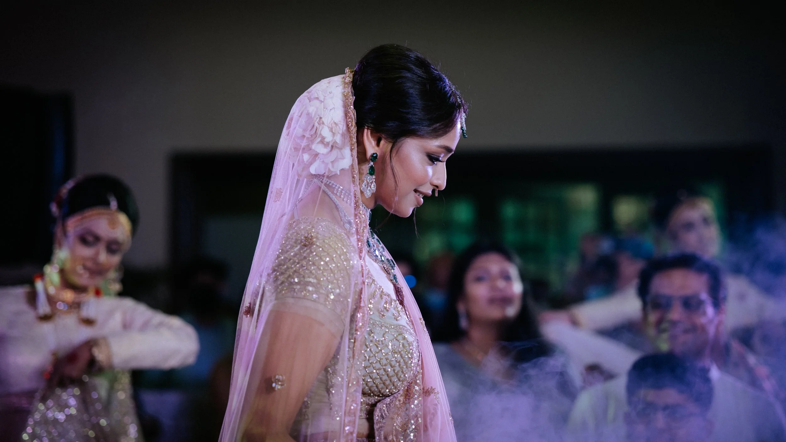 A woman dressed in traditional Indian attire, with ornate jewelry and a pink and gold sari, is smiling and looking down at a wedding or celebration event. Other women and guests are visible in the background.
