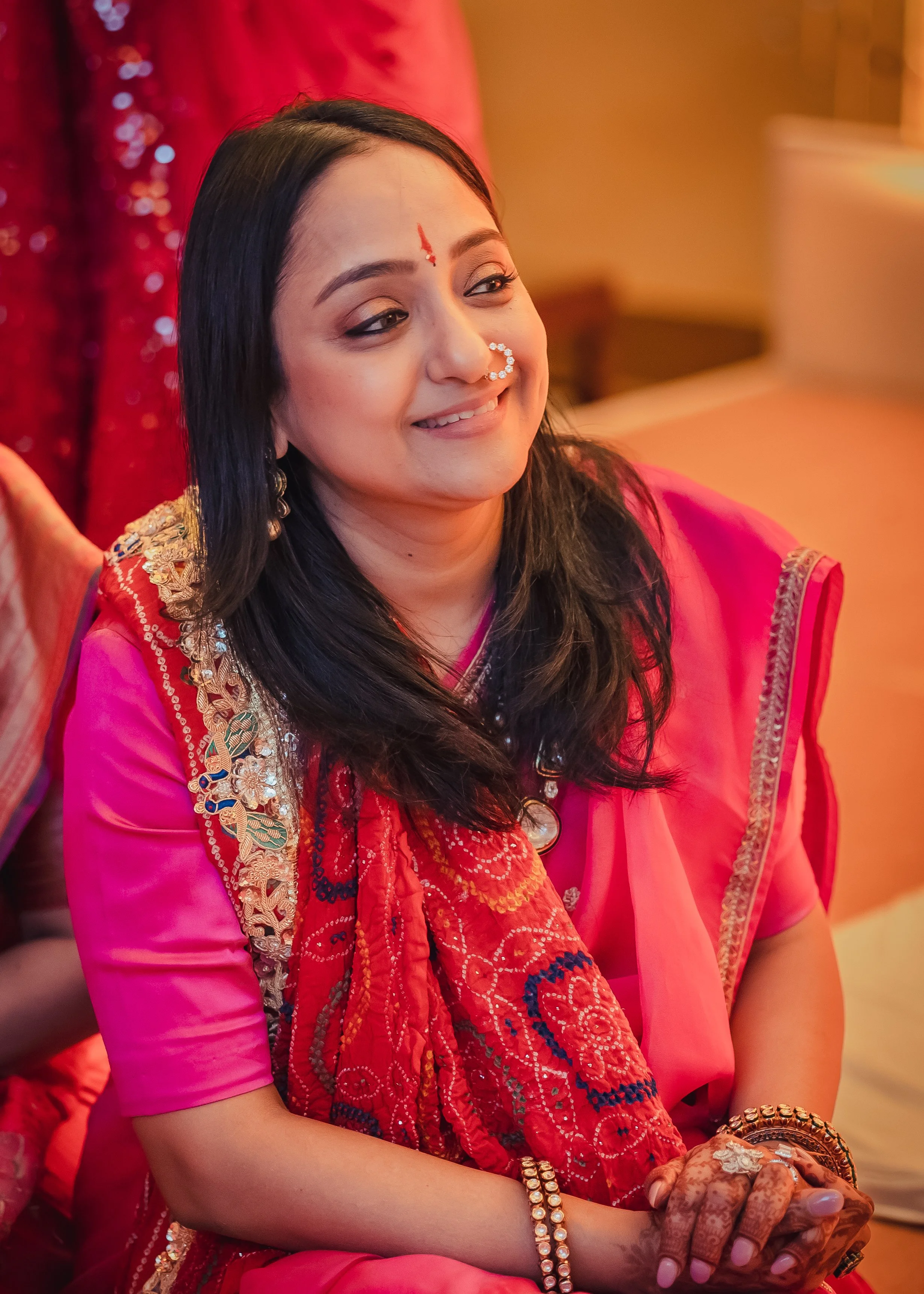 A woman in traditional Indian attire, wearing a pink sari with intricate embroidery and jewelry, smiling warmly at a celebration or event.