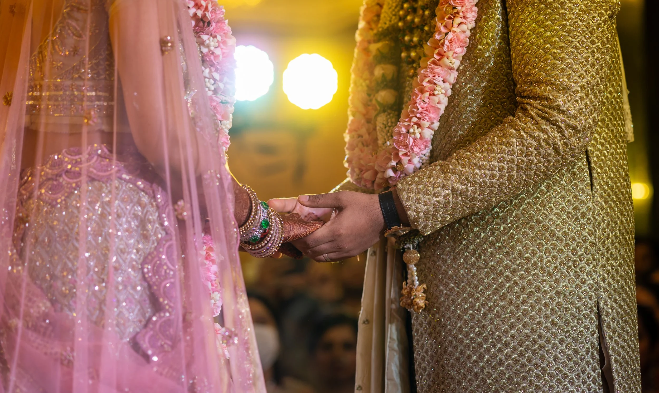 A groom holding the bride's hand during a traditional wedding ceremony, both wearing ornate clothing and floral garlands.
