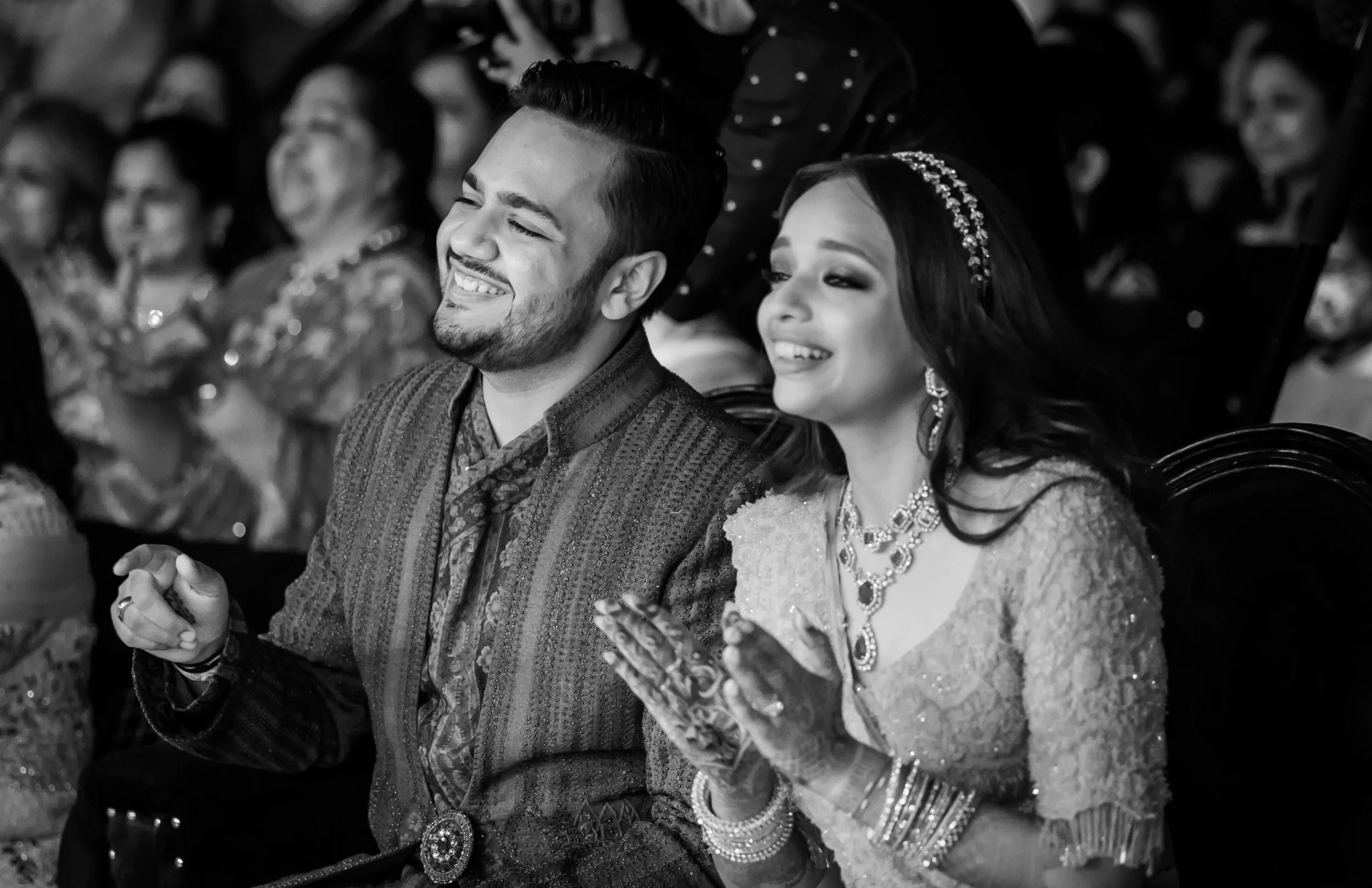 A smiling couple dressed in traditional Indian attire sitting in a crowd at a celebration or ceremony.