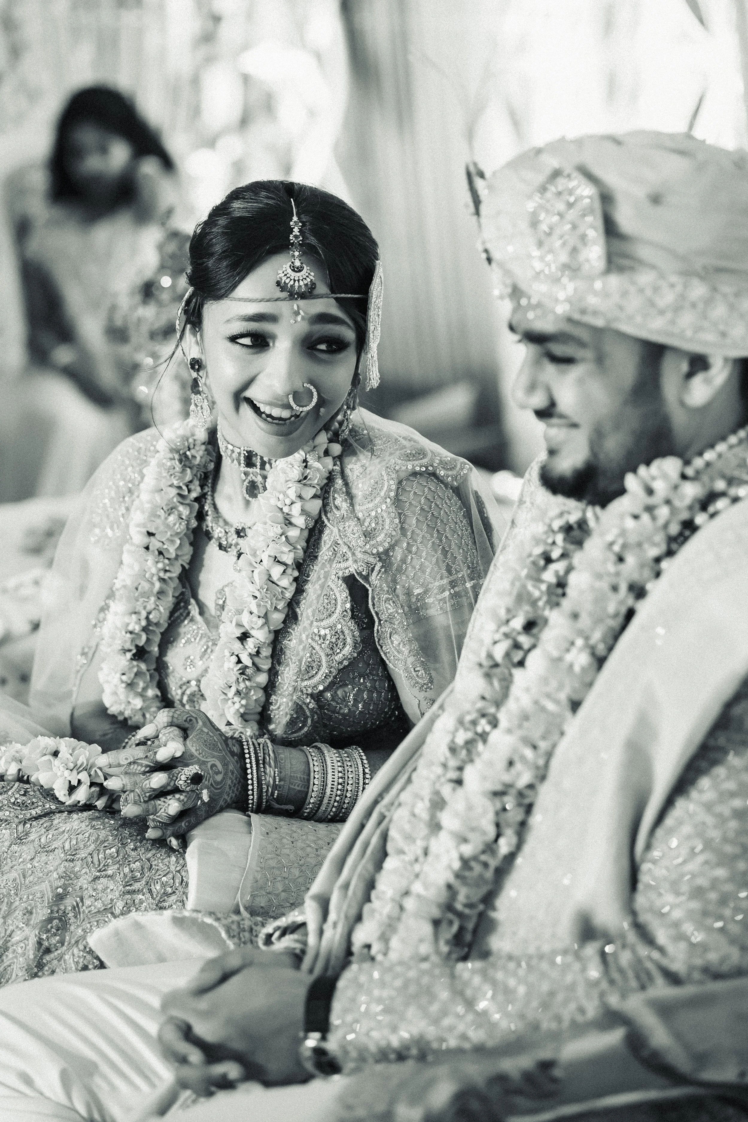 Black and white photograph of a bride and groom during a traditional Indian wedding ceremony, smiling and holding hands.