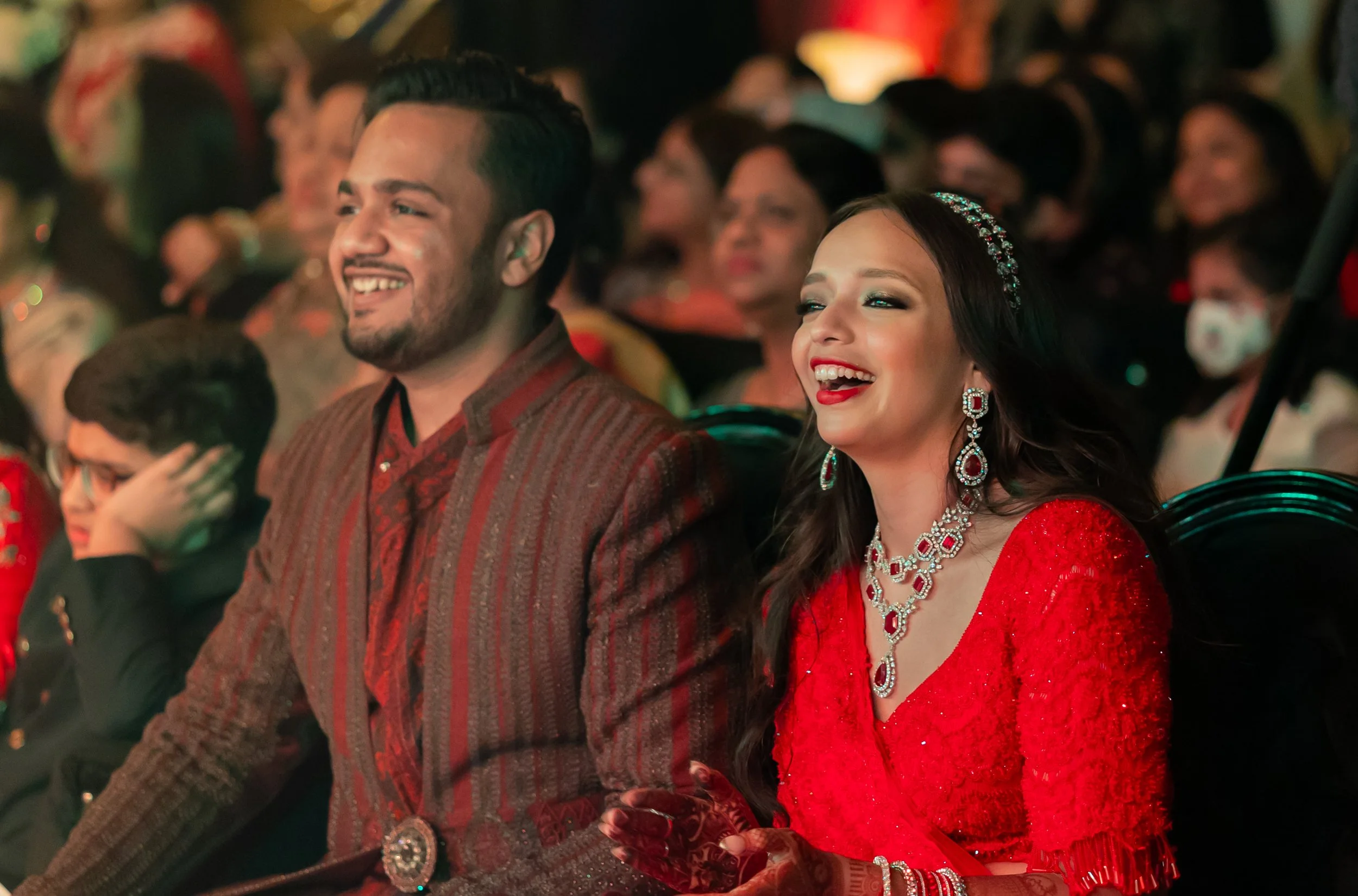 Smiling man and woman dressed in traditional South Asian clothing, sitting in an audience at a celebration or wedding, with other attendees in the background.